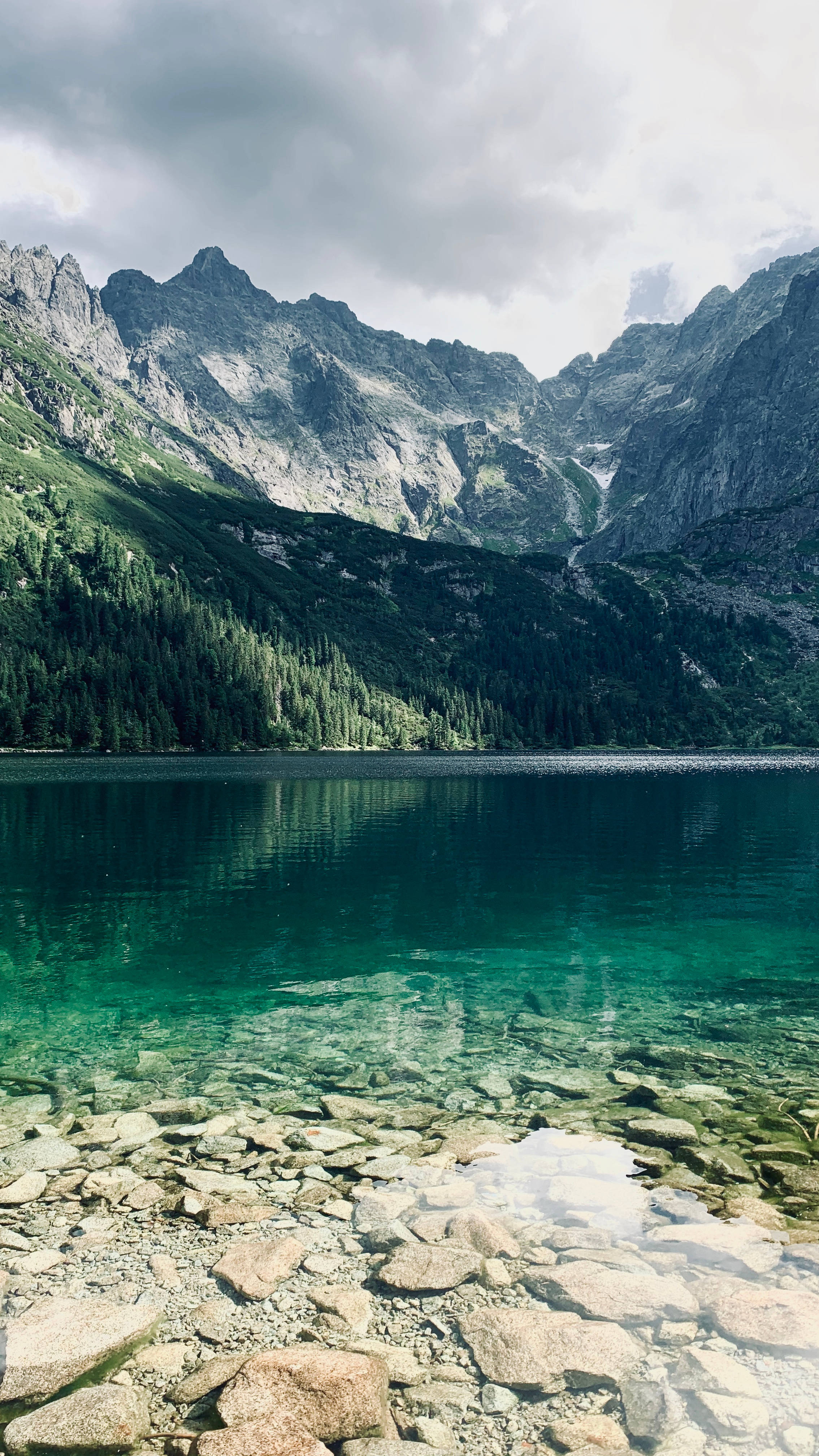 Lake 桌布 壁紙 — 一個寧靜的山間湖泊，清澈的碧綠湖水映照著崎嶇的山峰和茂密的常綠森林，天空多雲。