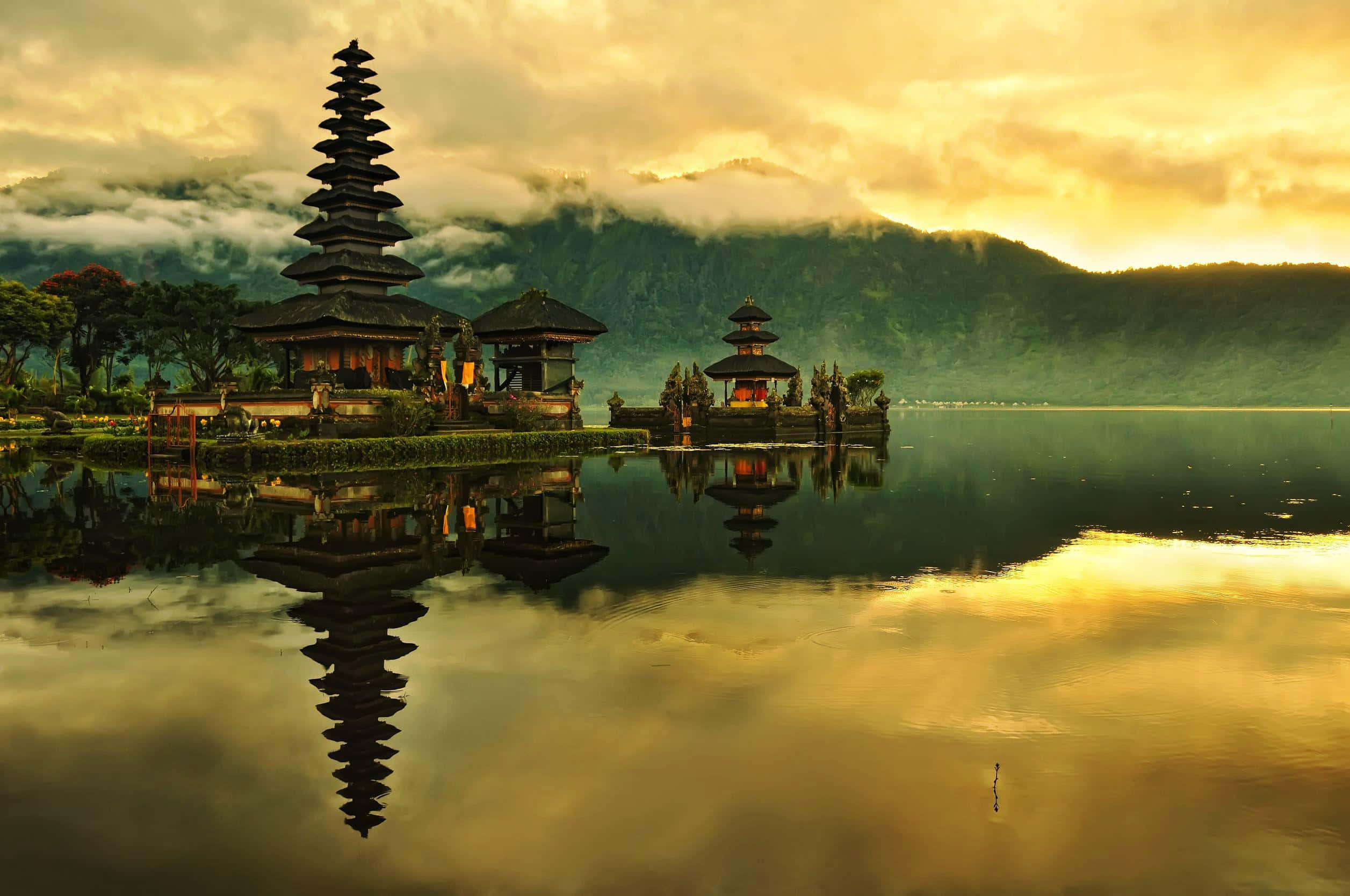 Bali Beach 桌布 壁紙 — 一個寧靜的峇里寺廟群完美地倒映在平靜的水面上，金色的晨光將霧氣繚繞的山巒沐浴在溫暖的色調中。