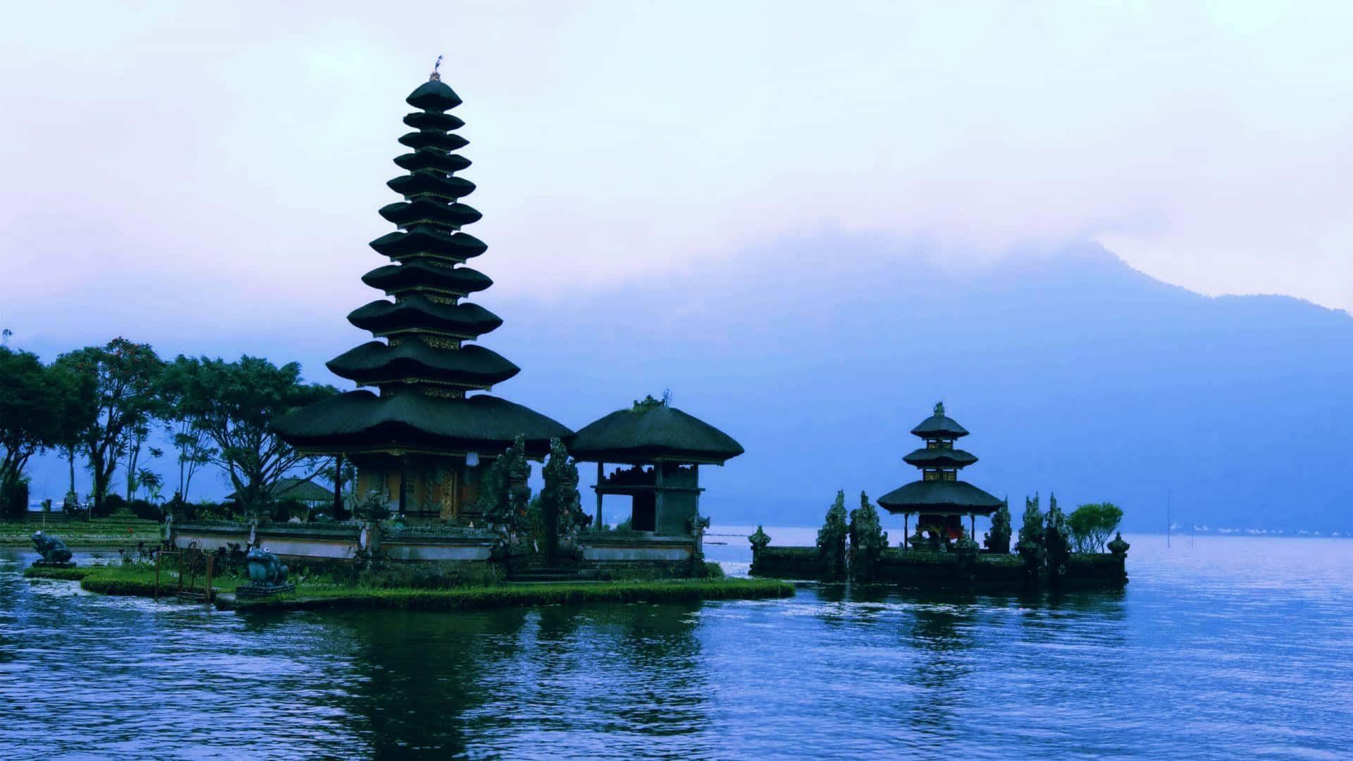 Bali Beach 桌布 壁紙 — 一個寧靜的峇里島寺廟建築群，層疊屋頂漂浮在平靜的藍色水面上，籠罩在霧氣朦朧的暮色天空下。