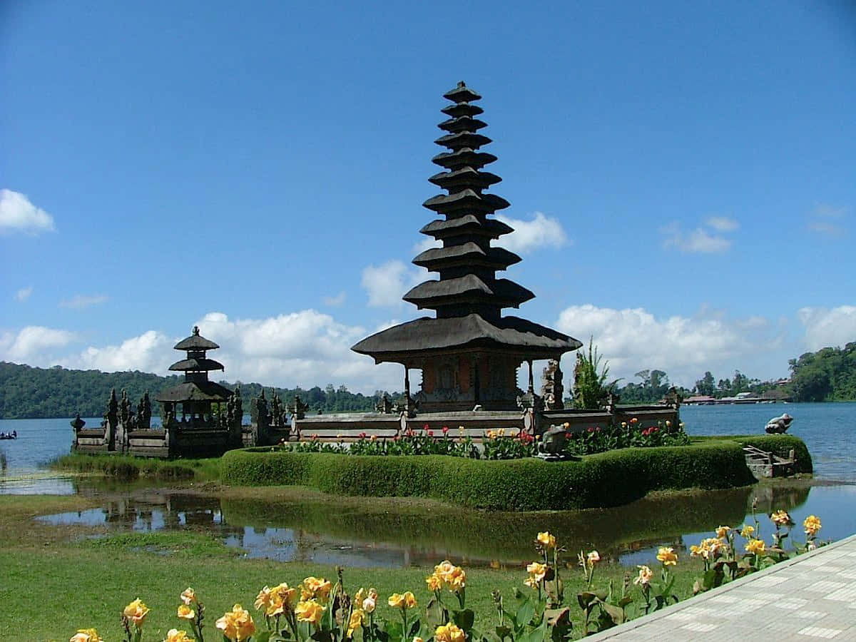 Bali 桌布 壁紙 — 一個寧靜的峇里島寺廟群，擁有層疊的寶塔，矗立在一個鬱鬱蔥蔥的綠色島嶼上，周圍環繞著平靜的藍色水域和盛開的黃色花朵，天空晴朗。