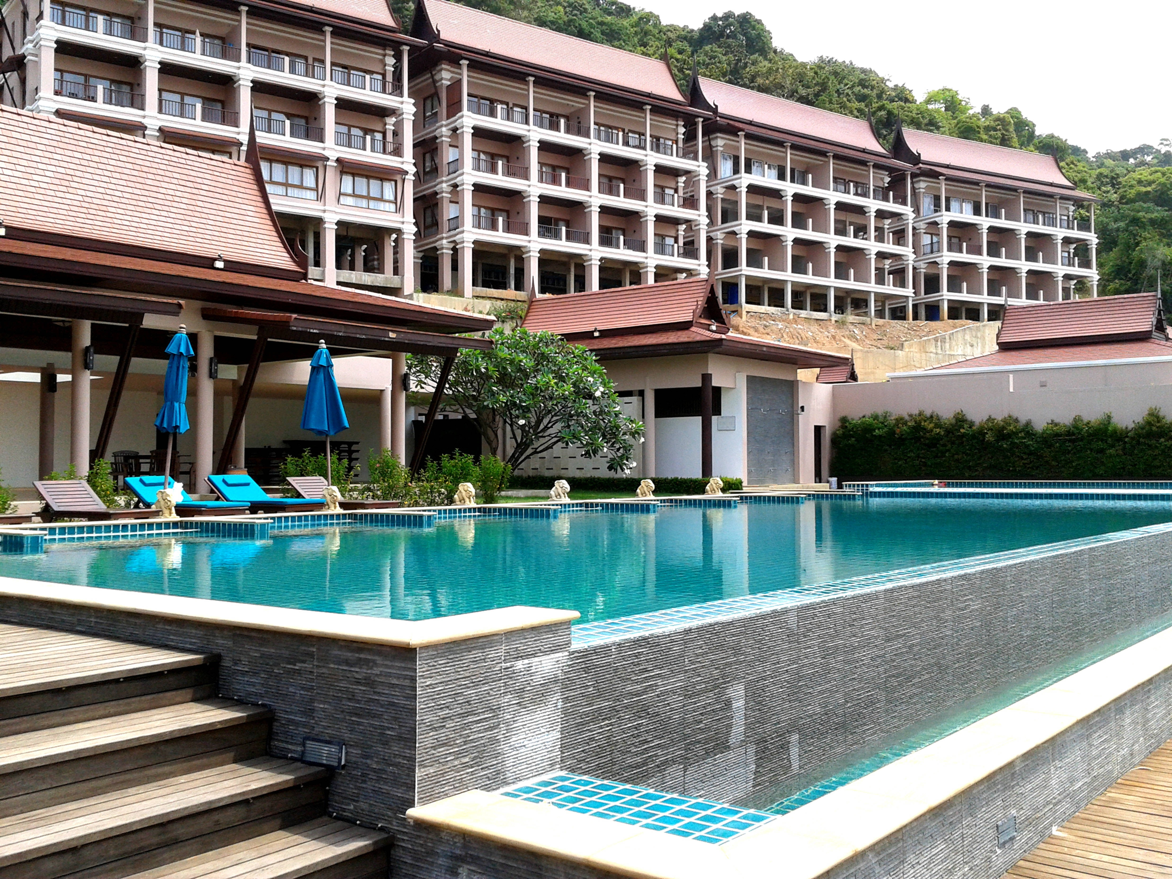 Hotel 桌布 壁紙 — 一個寧靜的度假村泳池，碧綠色的水在明亮的日光下映照著多層建築和鬱鬱蔥蔥的綠色山坡。