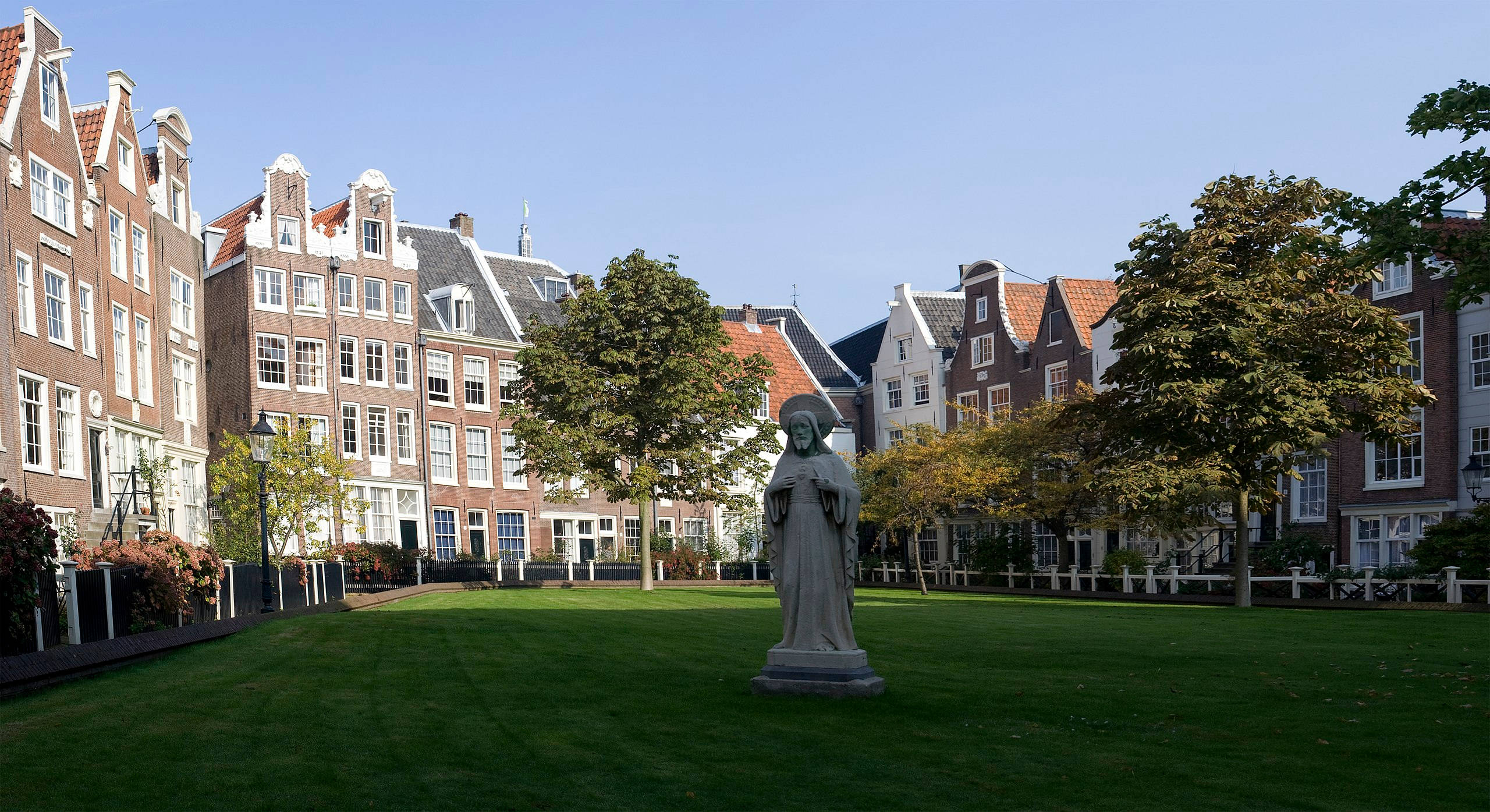 Amsterdam 桌布 壁紙 — 一個寧靜的庭院，中央有一座石像，立於茂密的綠草地上，周圍是經典的荷蘭山牆房屋，上方是清澈的藍天。