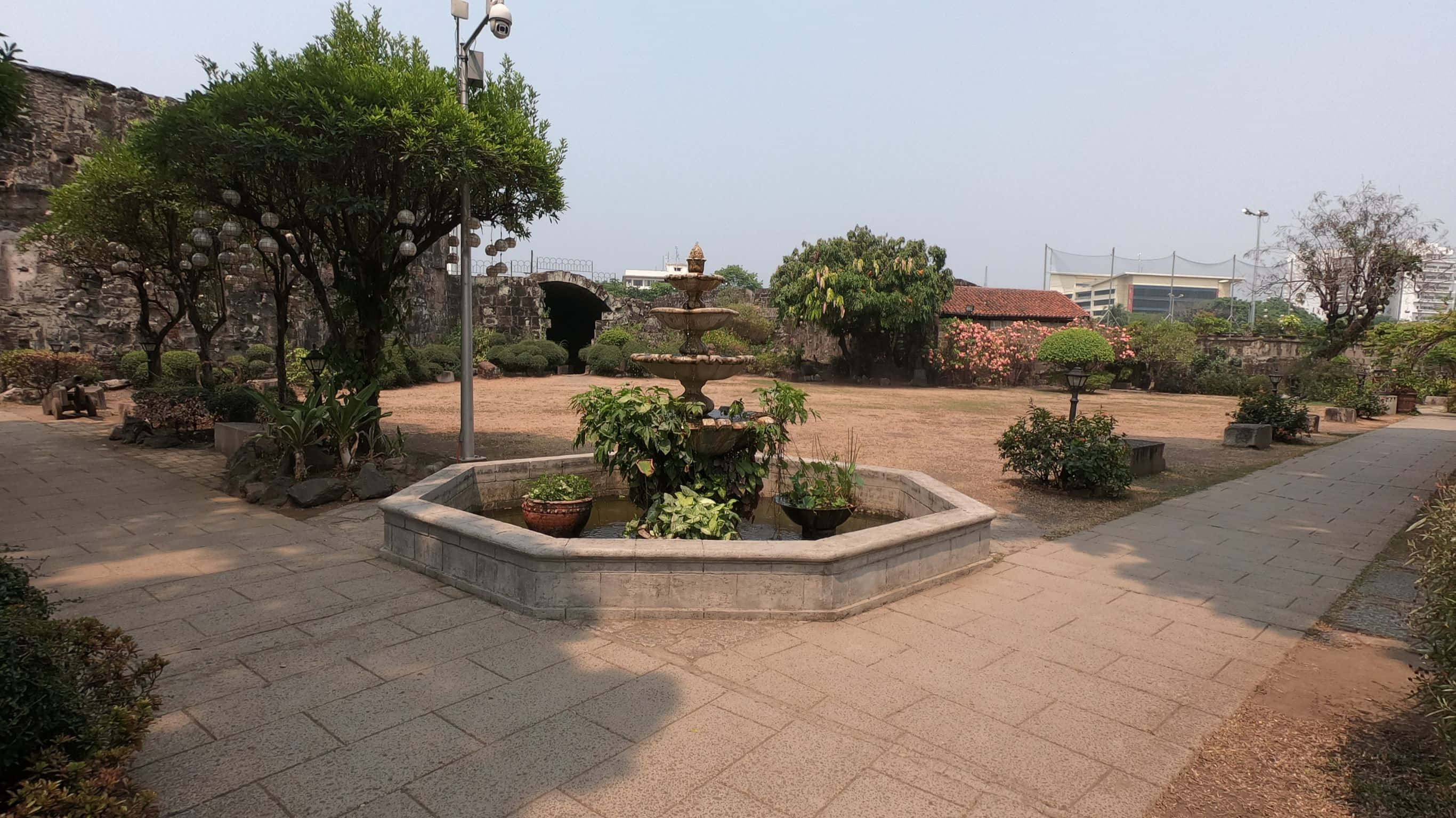 2732x1536 桌布 壁紙 — 一個寧靜的庭院，設有分層石噴泉，周圍是鋪砌的小徑和晴朗天空下的茂密綠植。
