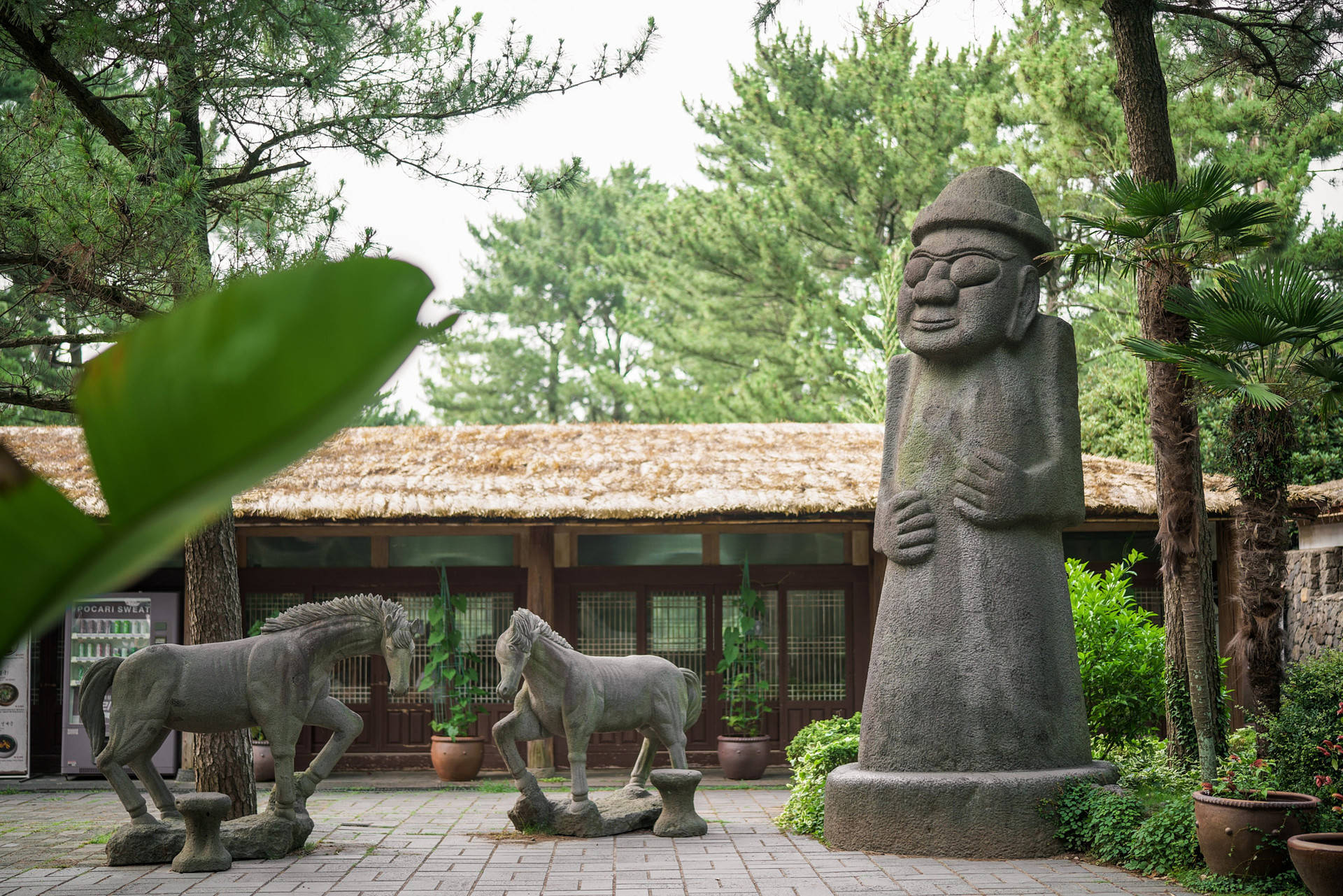 Jeju 桌布 壁紙 — 一個寧靜的庭院中，有一座微笑人物的高聳石像，旁邊有兩匹馬的雕塑，周圍是茂密的綠樹。