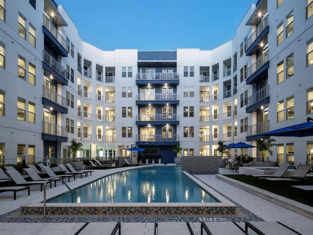 Building 桌布 壁紙 — 一個寧靜的庭院游泳池，周圍環繞著現代白色公寓樓，帶有藍色點綴和黃昏時發光的窗戶。