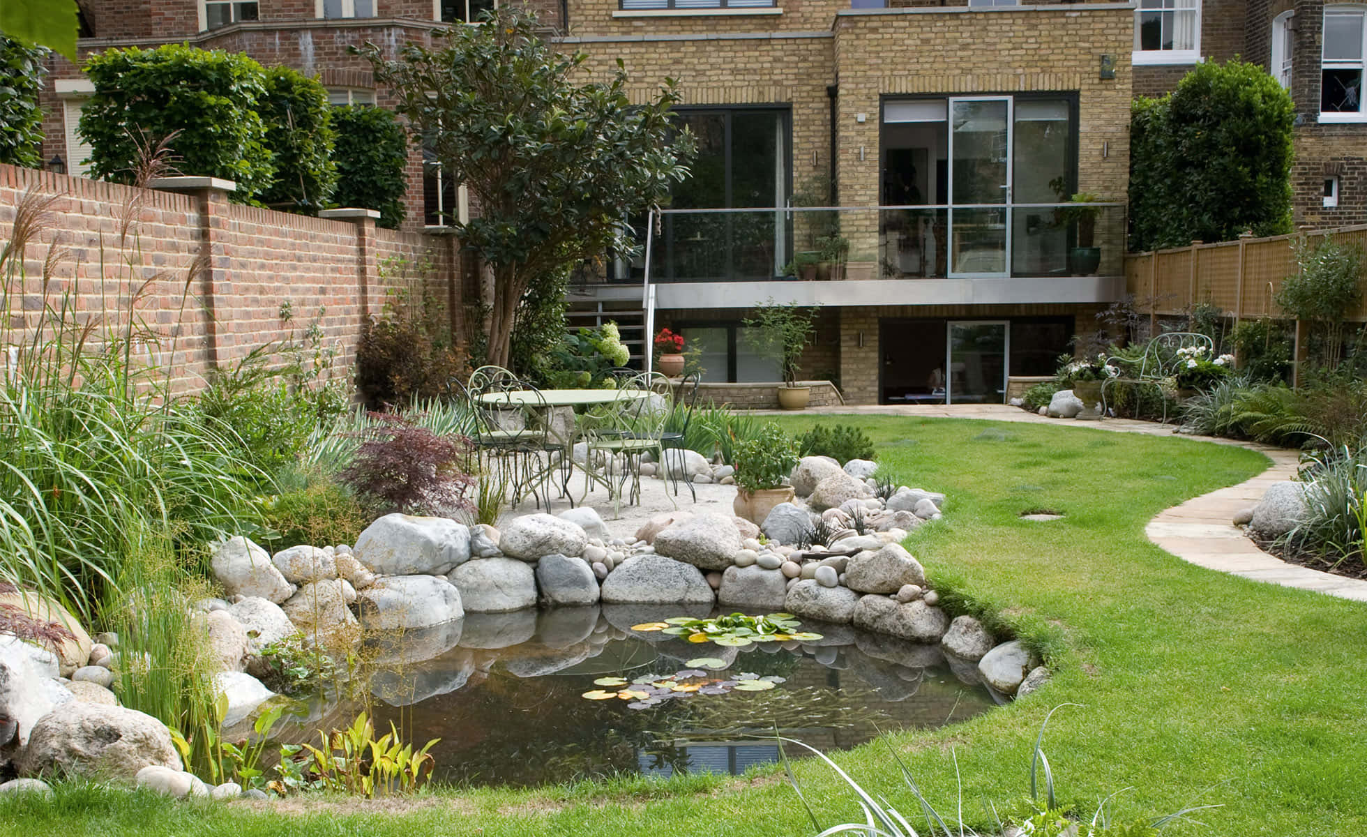 Garden Design 桌布 壁紙 — 一個寧靜的後院花園，有石邊池塘、睡蓮、綠色草坪和背景中的現代房屋。