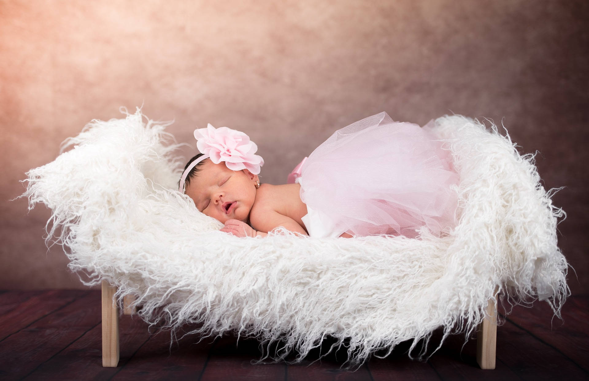 Very Cute Baby 桌布 壁紙 — 一個寧靜的新生女嬰穿著粉紅色芭蕾舞裙和花卉頭帶，在溫暖漸變背景前的蓬鬆白色床上安詳入睡。