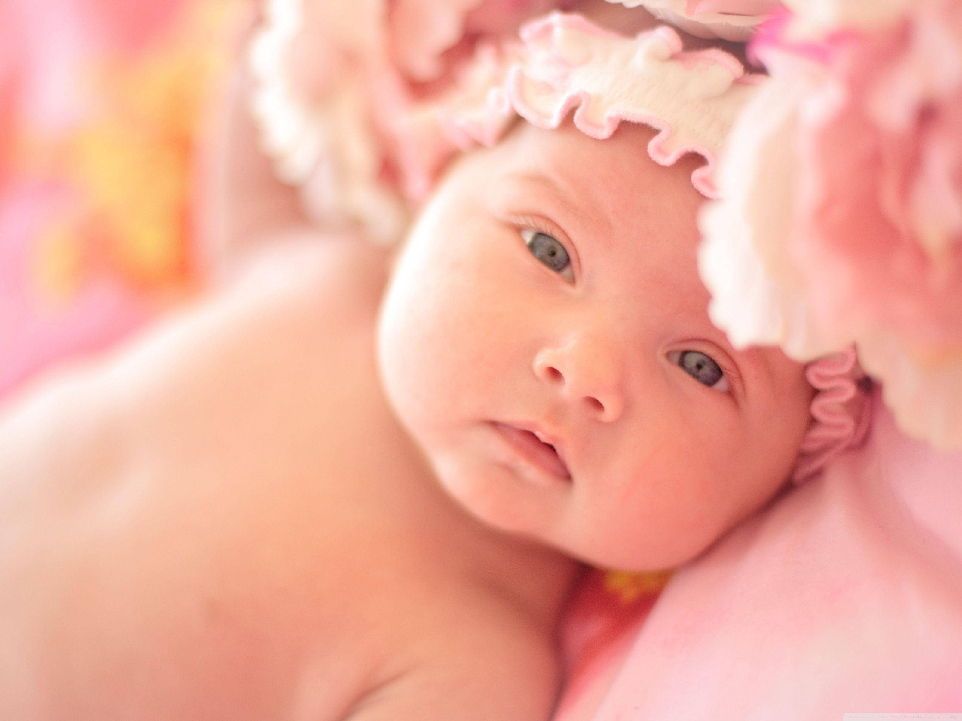 Newborn 桌布 壁紙 — 一個寧靜的新生嬰兒，有著大大的藍眼睛，戴著柔軟的粉紅色花卉帽，在夢幻般的粉彩背景中安詳地休息。