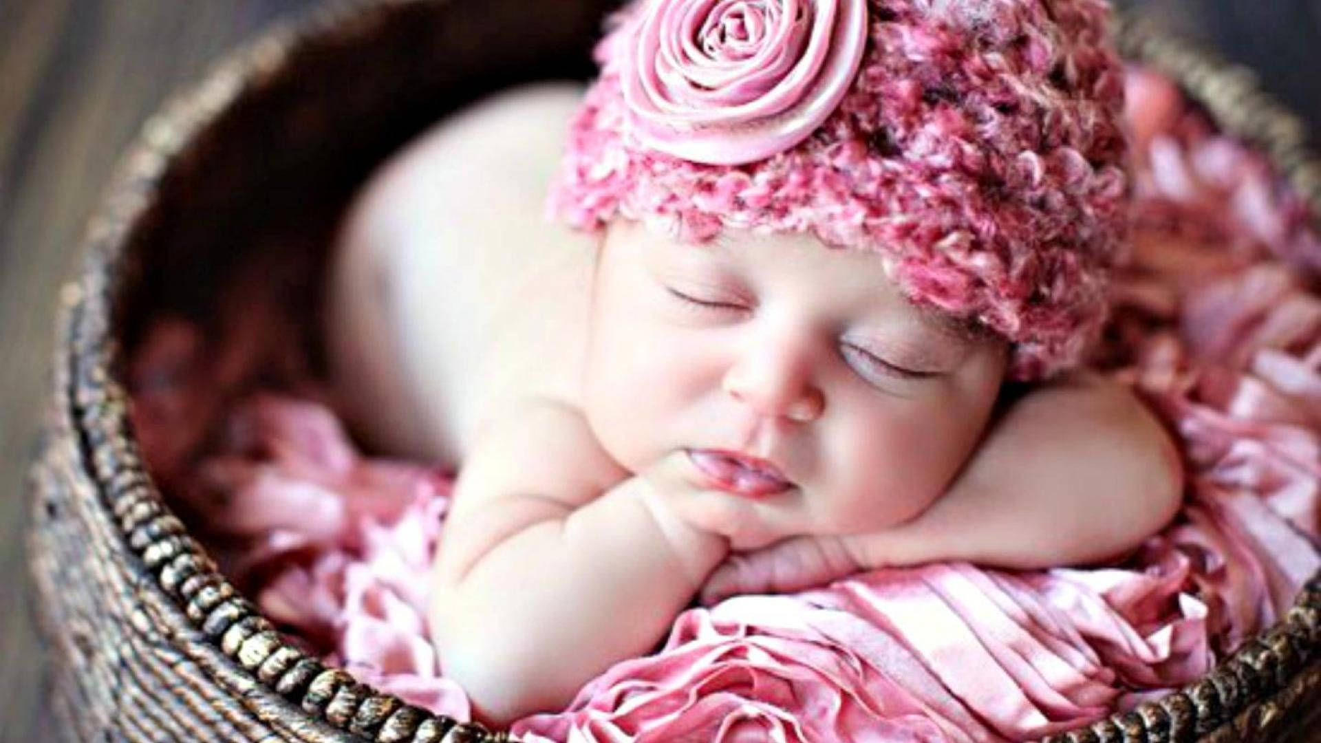 Newborn Baby 桌布 壁紙 — 一個寧靜的新生嬰兒在一個鋪有柔軟粉紅色布料、編織而成的籃子裡安詳地睡覺，戴著一頂帶有玫瑰花的粉紅色鉤針帽子。