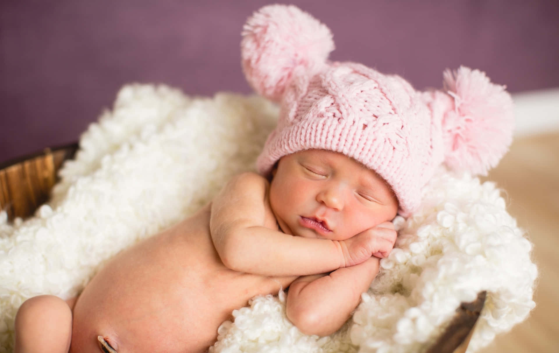 Infant 桌布 壁紙 — 一個寧靜的新生嬰兒在一個鋪滿蓬鬆白色羊毛的木籃裡安詳地睡覺，戴著一頂柔軟的粉紅色針織帽，上面有絨球。
