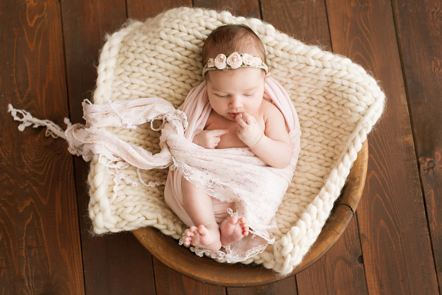 Newborn Baby 桌布 壁紙 — 一個寧靜的新生嬰兒在深色木地板上的一個木碗裡，被柔軟的粉紅色蕾絲和厚實的針織毯包裹著，安詳地睡著。