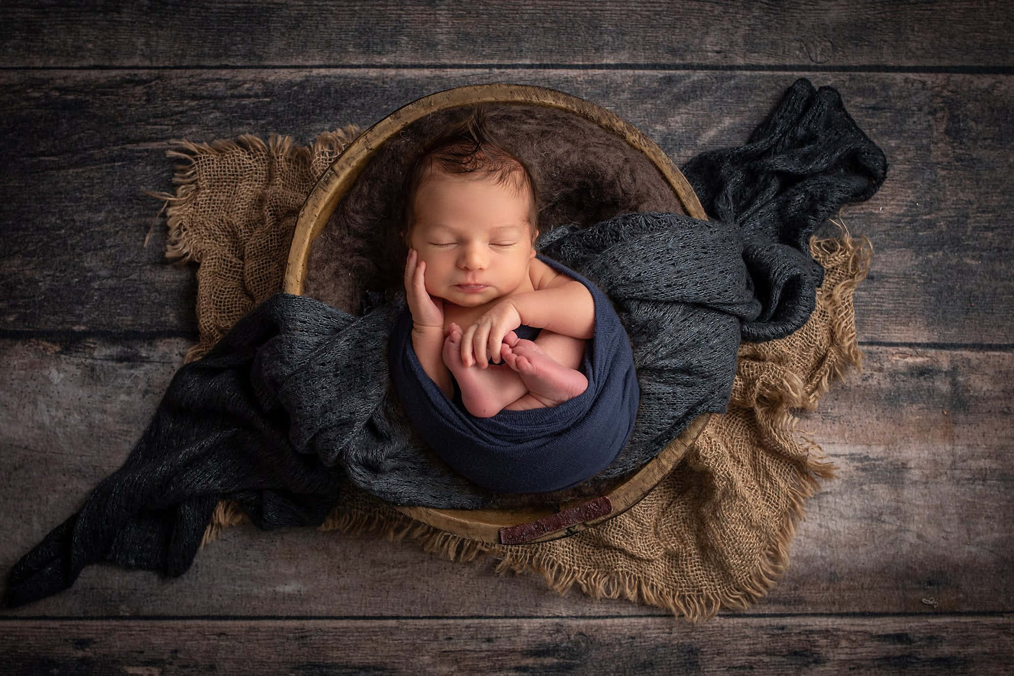 Newborn Baby 桌布 壁紙 — 一個寧靜的新生嬰兒在粗糙的木地板上，用海軍藍布料包裹著，安詳地睡在一個質樸的編織籃子裡。