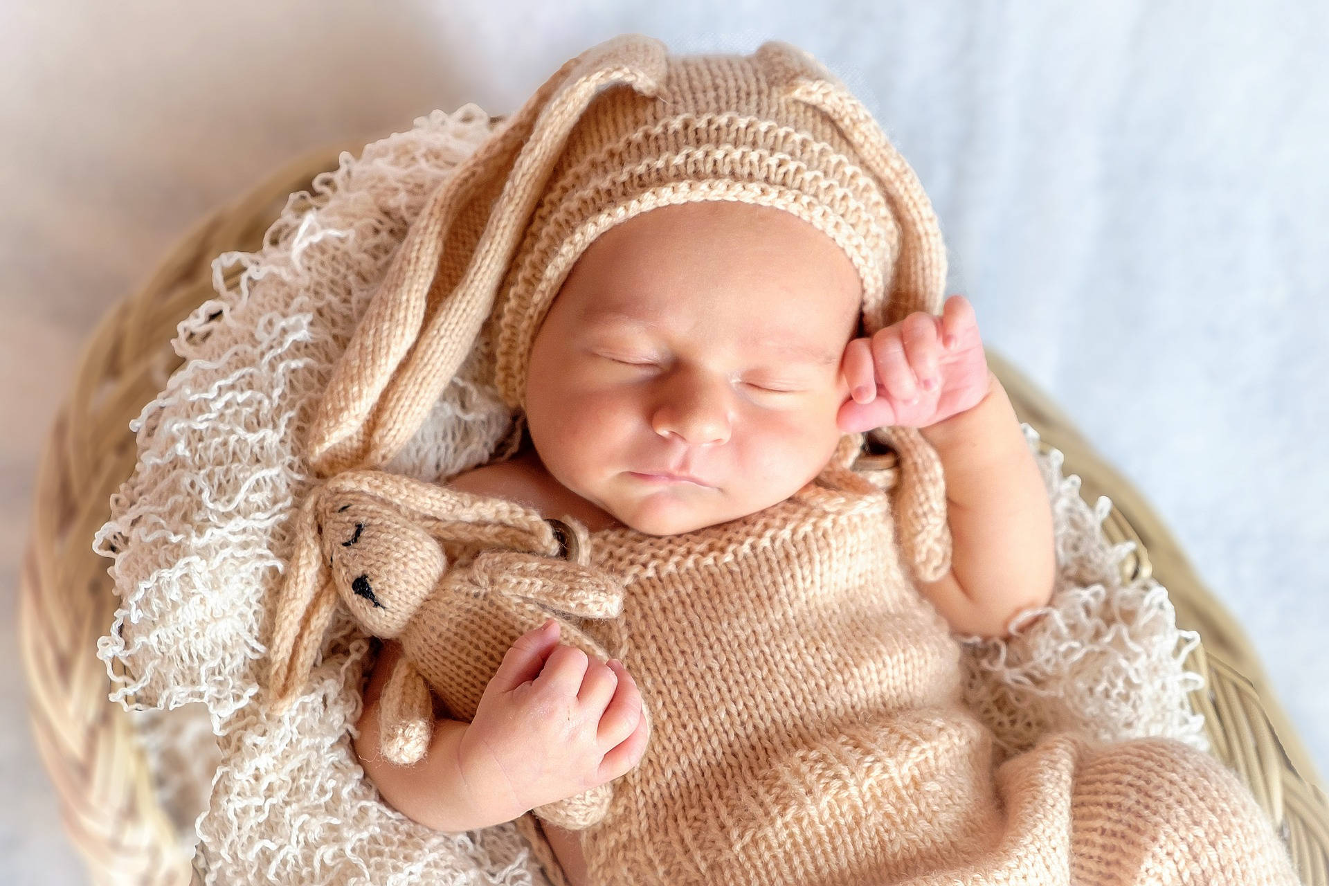 Very Cute Baby 桌布 壁紙 — 一個寧靜的新生嬰兒在編織籃中安詳入睡，包裹在柔軟的米色針織衣物中，並抱著一個相配的兔子毛絨玩具。