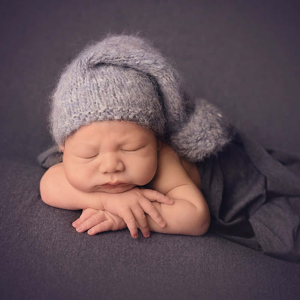 Newborn Baby 桌布 壁紙 — 一個寧靜的新生嬰兒戴著柔軟的灰色針織帽，帽上有毛茸茸的絨球，安睡在深灰色有紋理的毯子上。