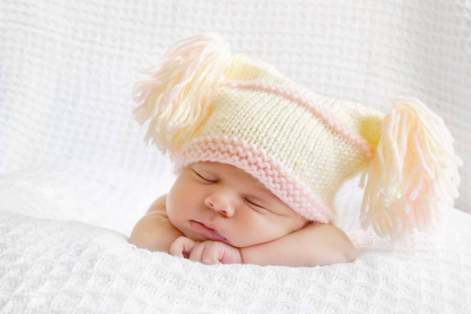 Baby Girl 桌布 壁紙 — 一個寧靜的新生嬰兒戴著柔軟的粉彩針織帽，帽上有毛絨絨的絨球，躺在有紋理的白色毛毯上安詳地睡覺。