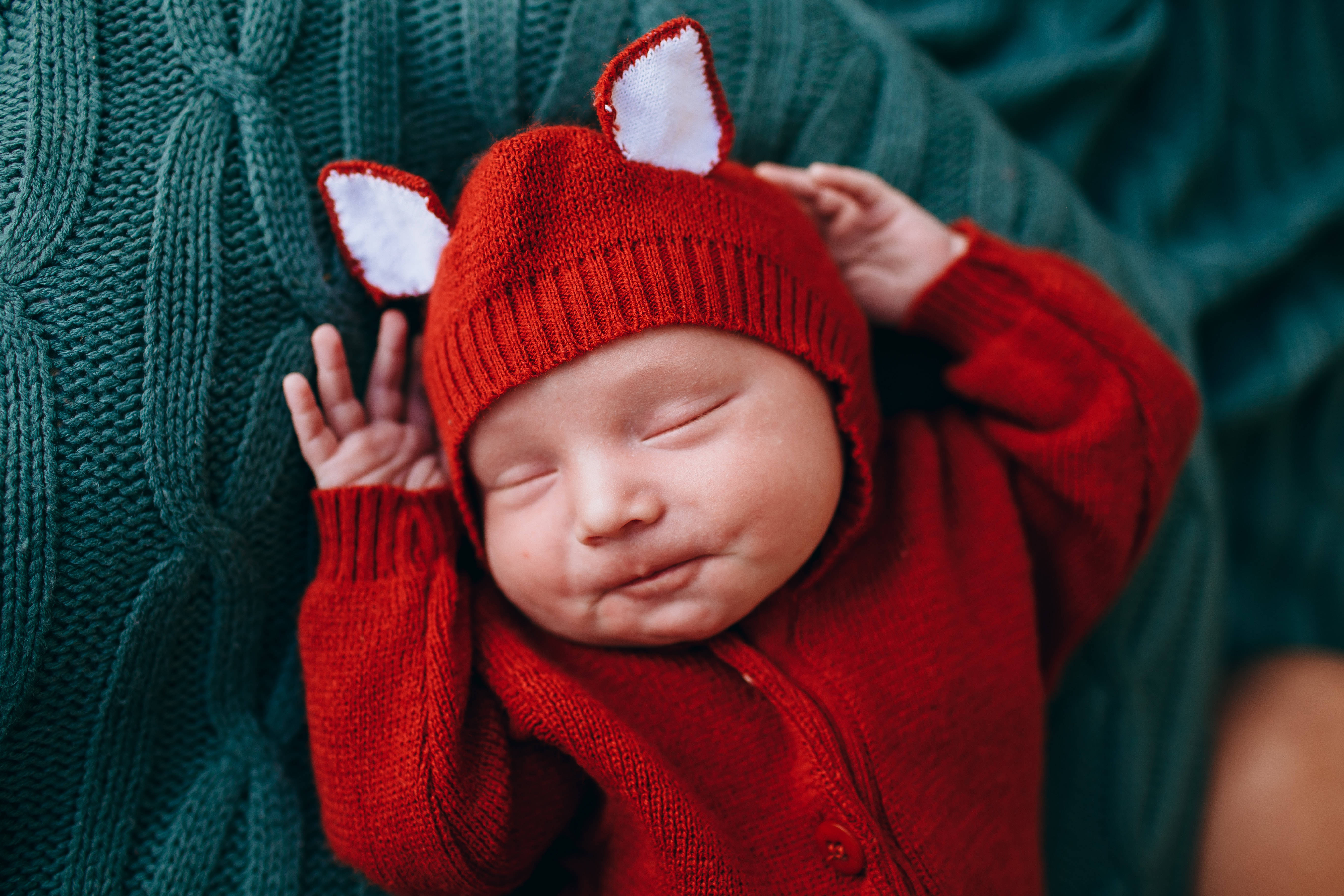 Very Cute Baby 桌布 壁紙 — 一個寧靜的新生嬰兒戴著紅色狐狸耳針織帽和配套服裝，在青綠色的電纜針織毯上安然入睡。