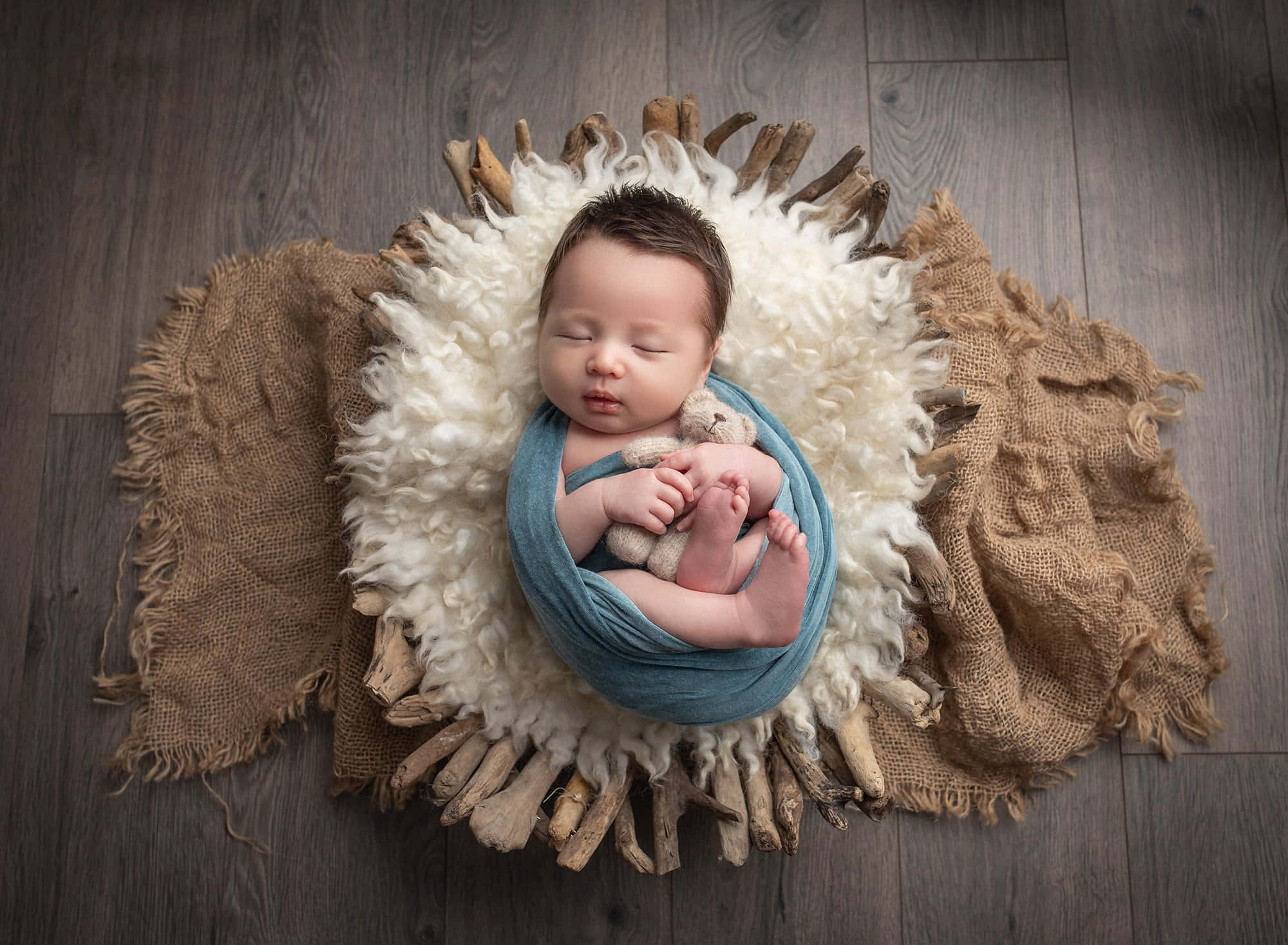 Newborn Baby 桌布 壁紙 — 一個寧靜的新生嬰兒用藍色布料包裹著，躺在木地板上的蓬鬆白色和黃麻巢中睡覺。
