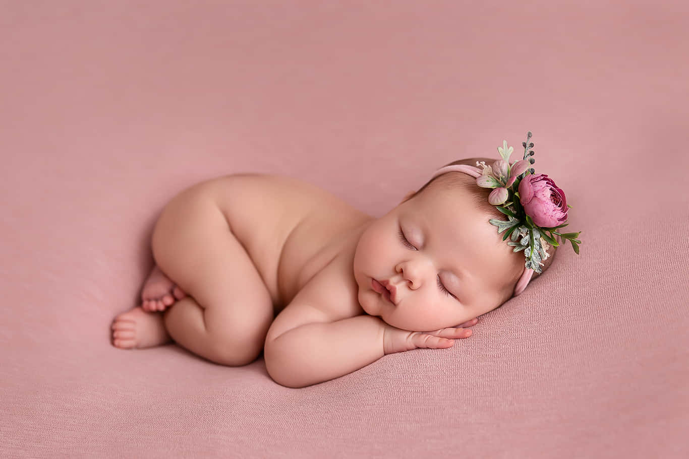Newborn Baby 桌布 壁紙 — 一個寧靜的新生嬰兒蜷縮在柔軟的粉紅色布料上睡覺，戴著精緻的花卉頭帶。