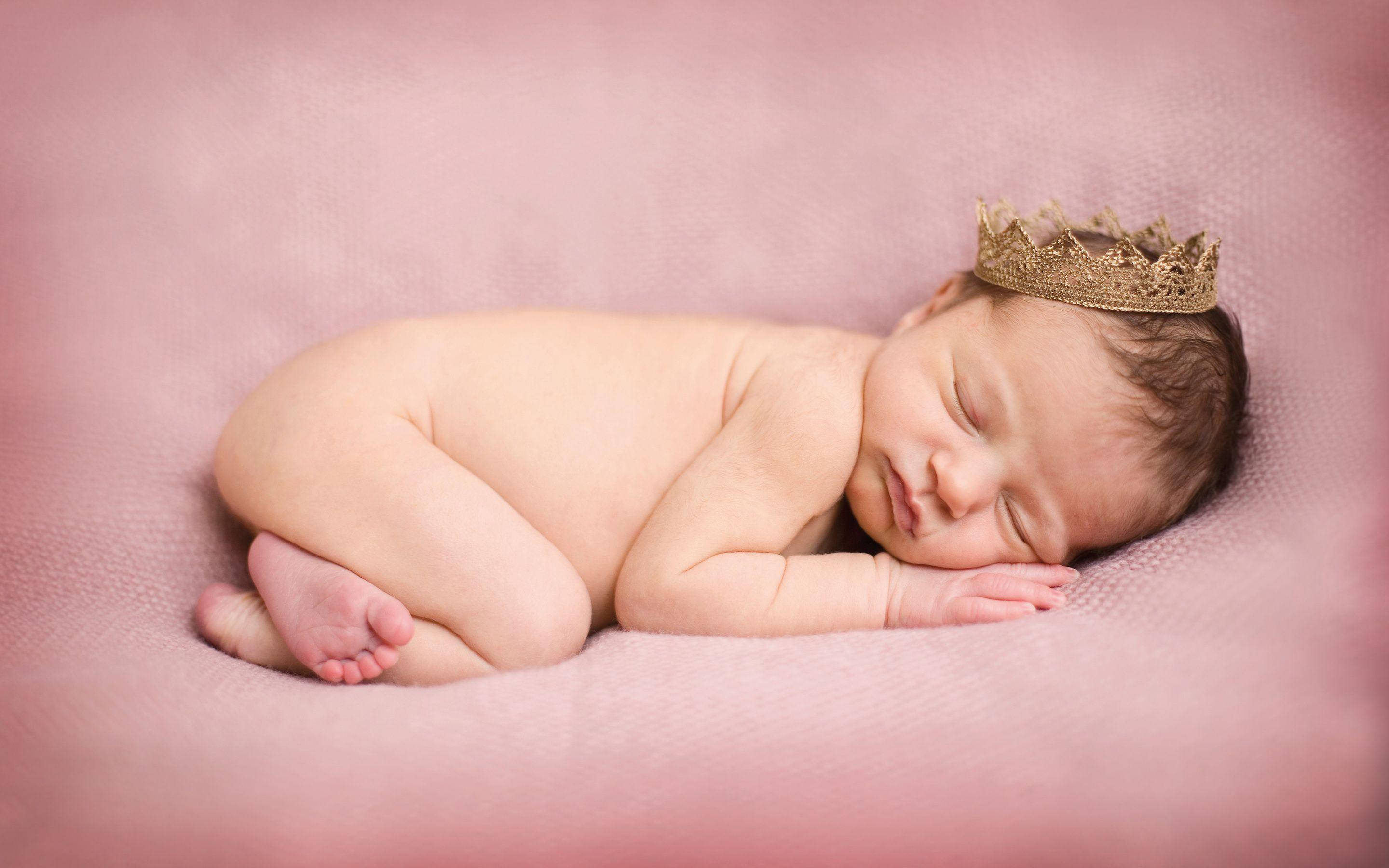 Newborn Baby 桌布 壁紙 — 一個寧靜的新生嬰兒蜷縮在柔軟的粉紅色紋理毛毯上睡覺，戴著精緻的金色皇冠。