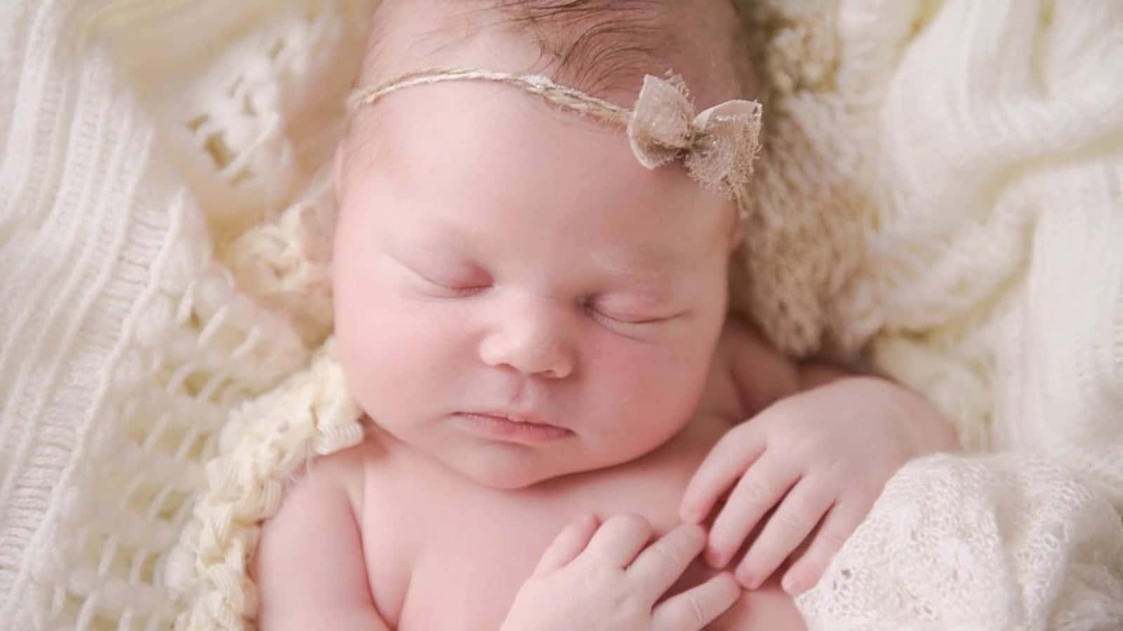 Baby Girl 桌布 壁紙 — 一個寧靜的新生嬰兒被柔軟的奶油色針織布料包裹著，戴著精緻的蝴蝶結頭帶，安詳地睡著。