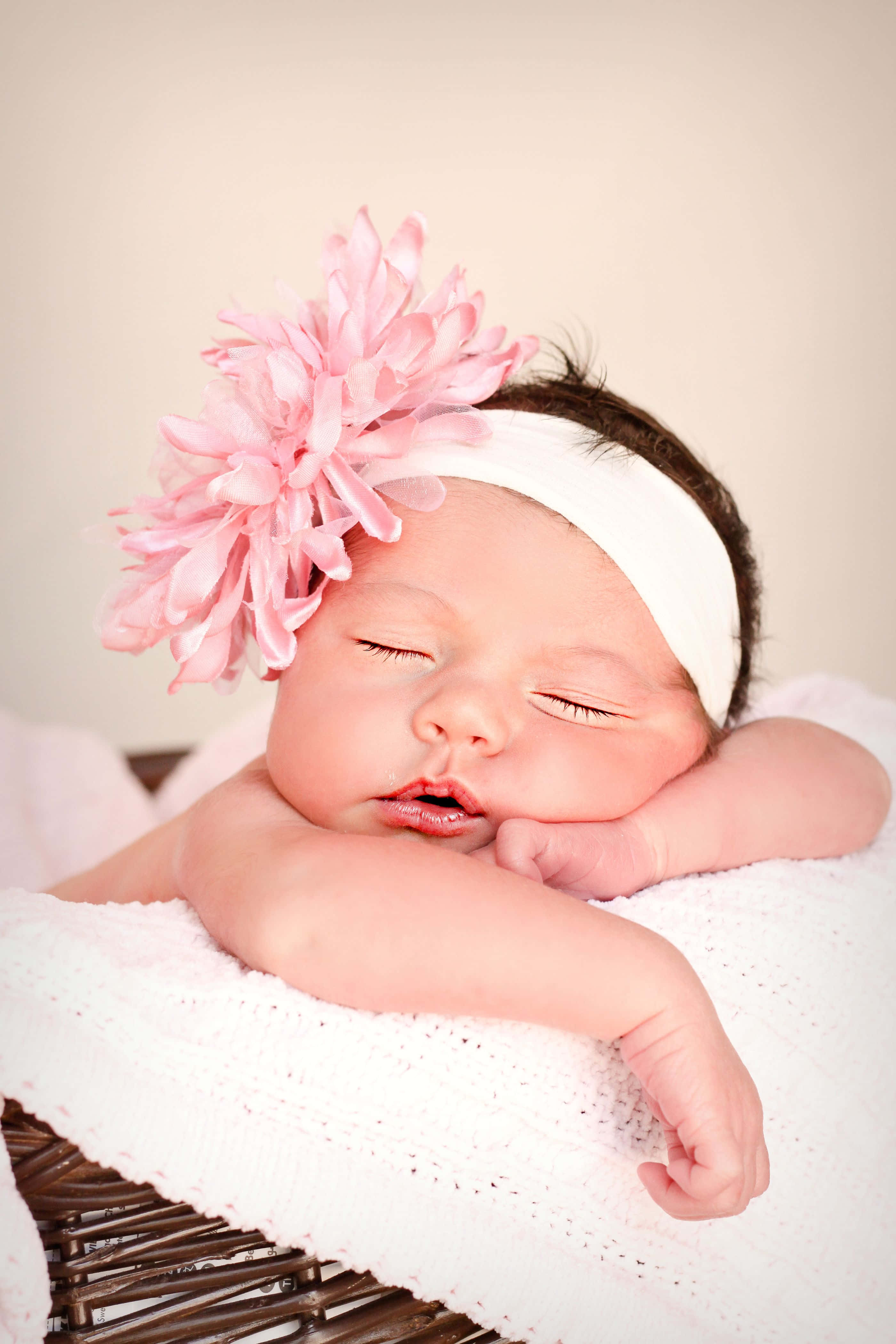 Baby Girl 桌布 壁紙 — 一個寧靜的新生嬰兒閉著眼睛安詳地睡覺，戴著一個飾有大粉紅色花朵的白色頭帶，包裹在白色針織毯子裡。