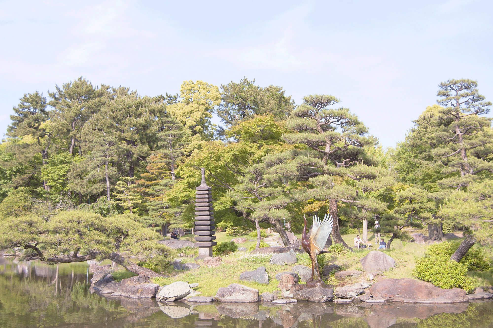 Nagoya 桌布 壁紙 — 一個寧靜的日本花園池塘倒映著石塔和鶴雕像，周圍環繞著茂密的綠樹，天空明亮。