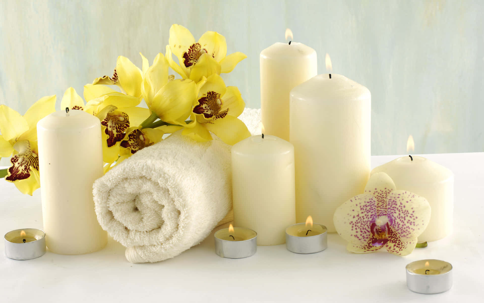 Spa Treatments 桌布 壁紙 — 一個寧靜的水療佈置，包含點燃的蠟燭、黃色蘭花和一條捲起的毛巾，置於白色表面上。