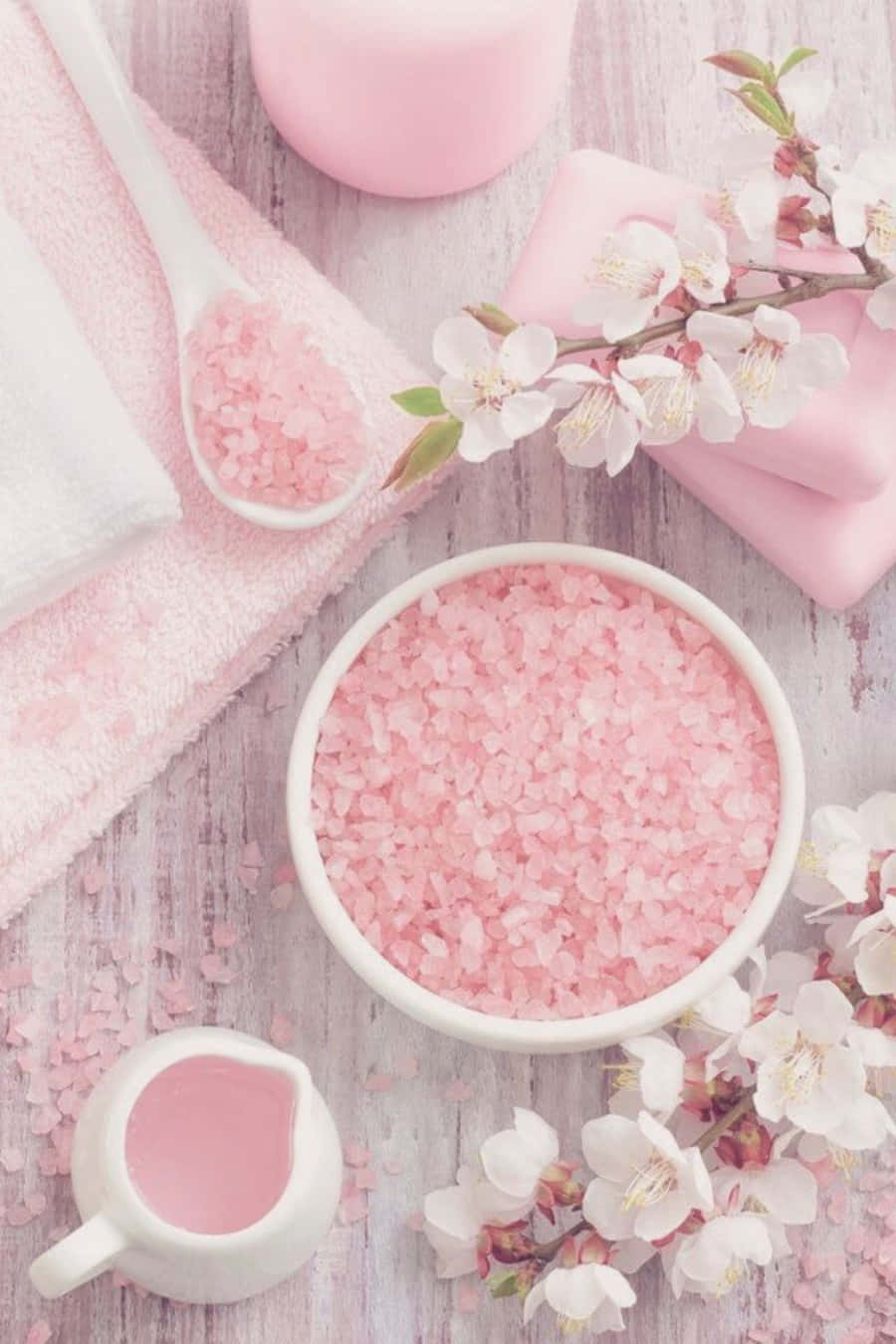 Pastel Pink Aesthetic 桌布 壁紙 — 一個寧靜的水療設置，粉紅鹽、花朵和毛巾擺放在柔軟的木質表面上。