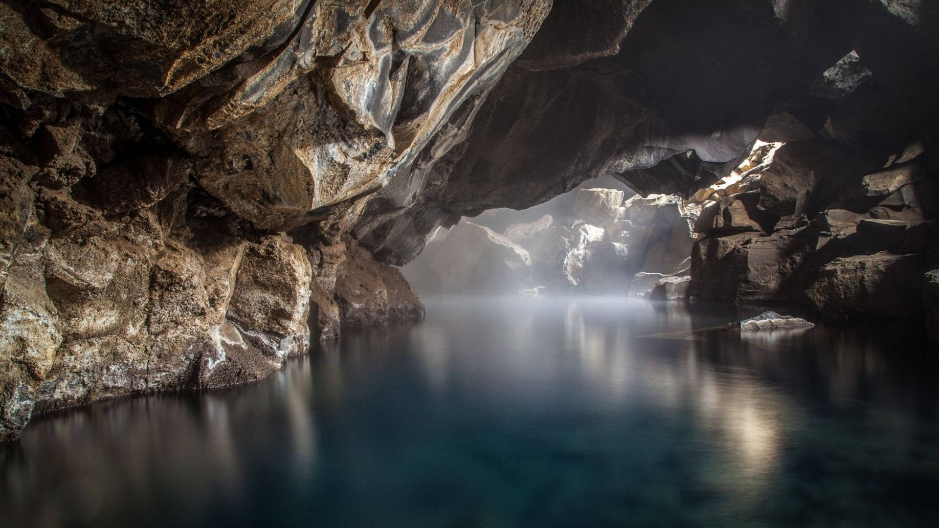 Cave 桌布 壁紙 — 一個寧靜的洞穴，平靜的藍色水面映照著崎嶇的岩石形態，柔和的光線從洞口滲入。