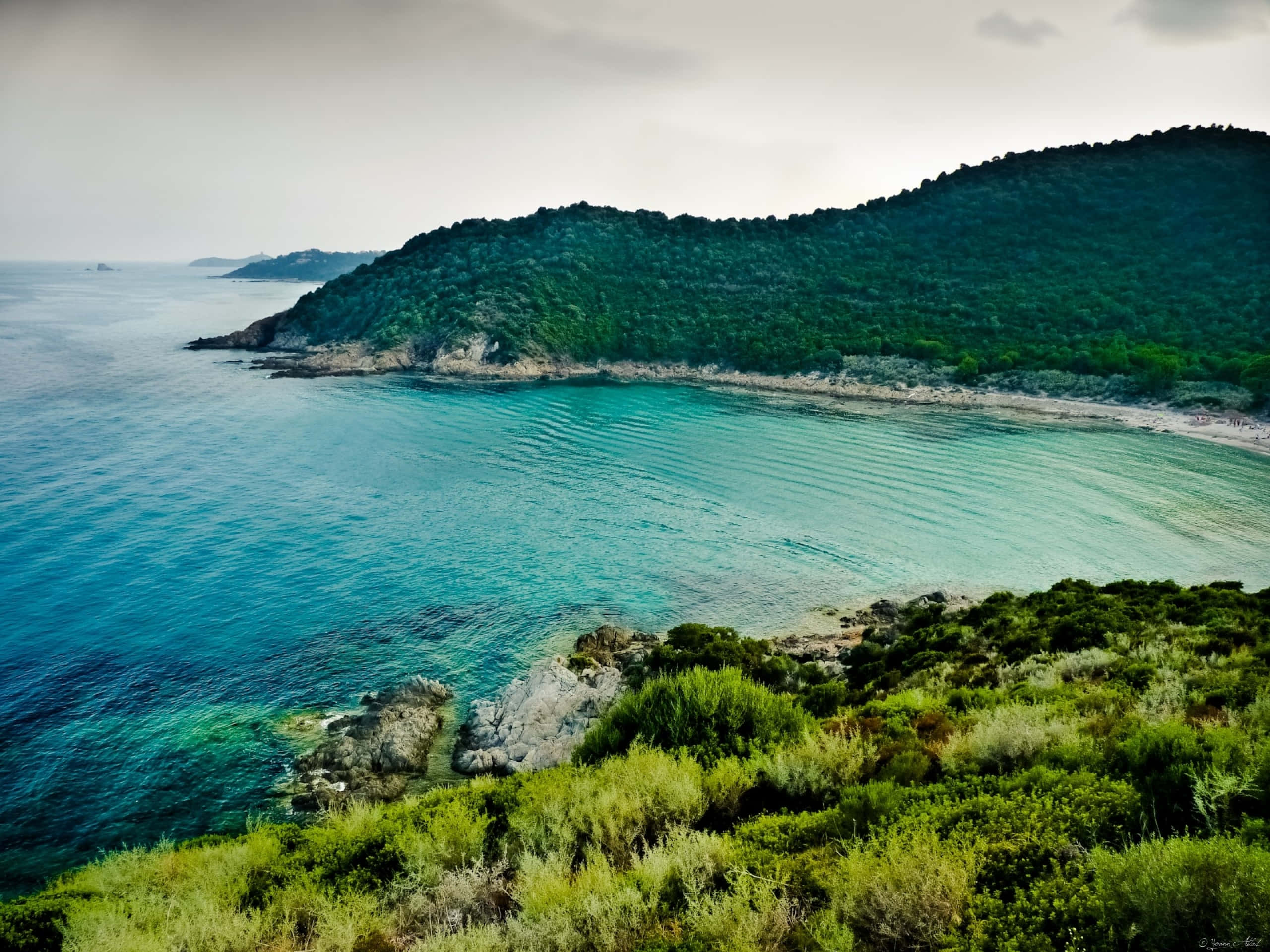 Beautiful Sea 桌布 壁紙 — 一個寧靜的海岸海灣，碧綠的海水輕輕拍打著岩石海岸，兩旁是鬱鬱蔥蔥的綠色山丘，天空多雲。