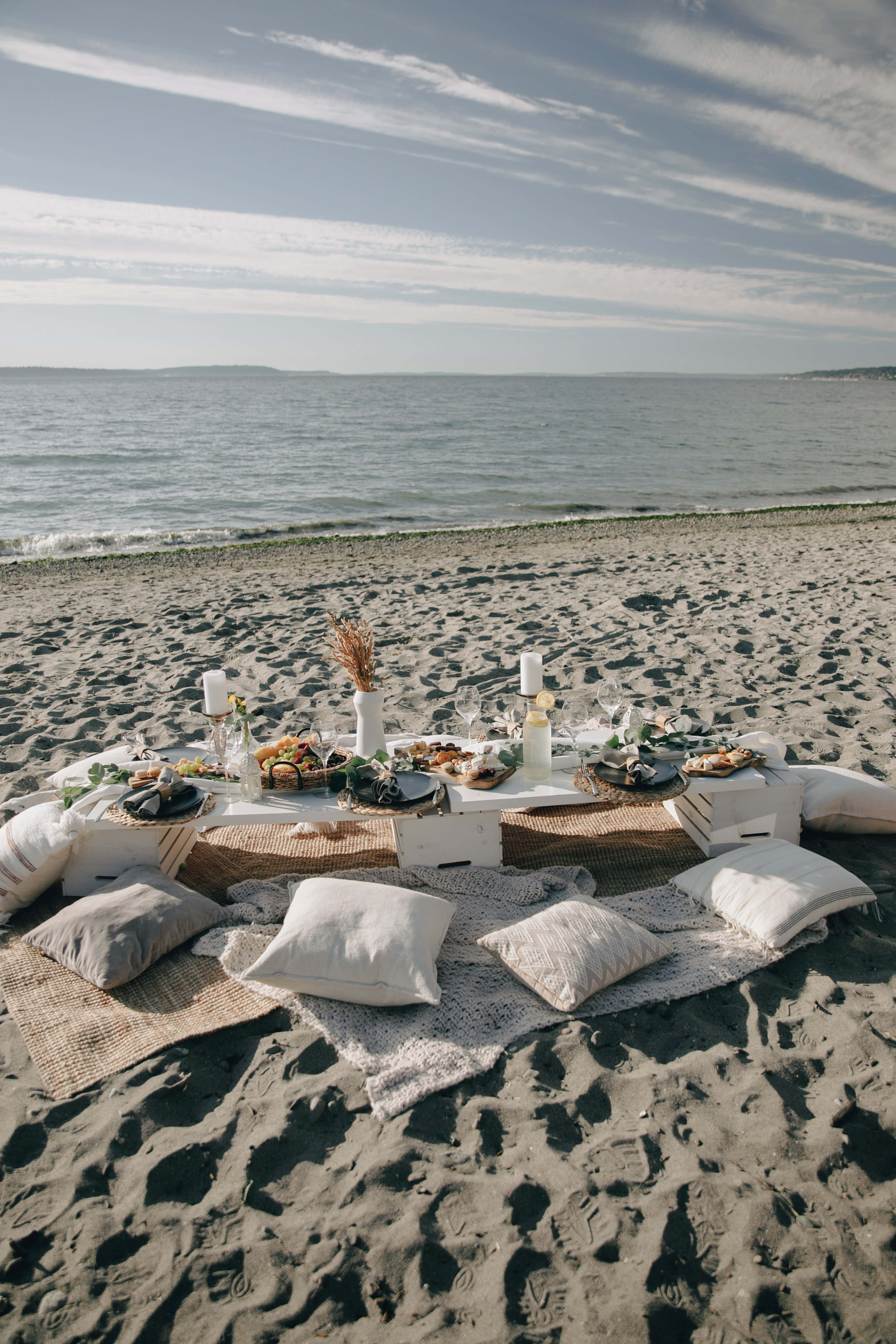 Beach Picnic 桌布 壁紙 — 一個寧靜的海灘野餐佈置，配有白色桌子、靠墊和食物，在明亮的天空和散落的雲朵下。