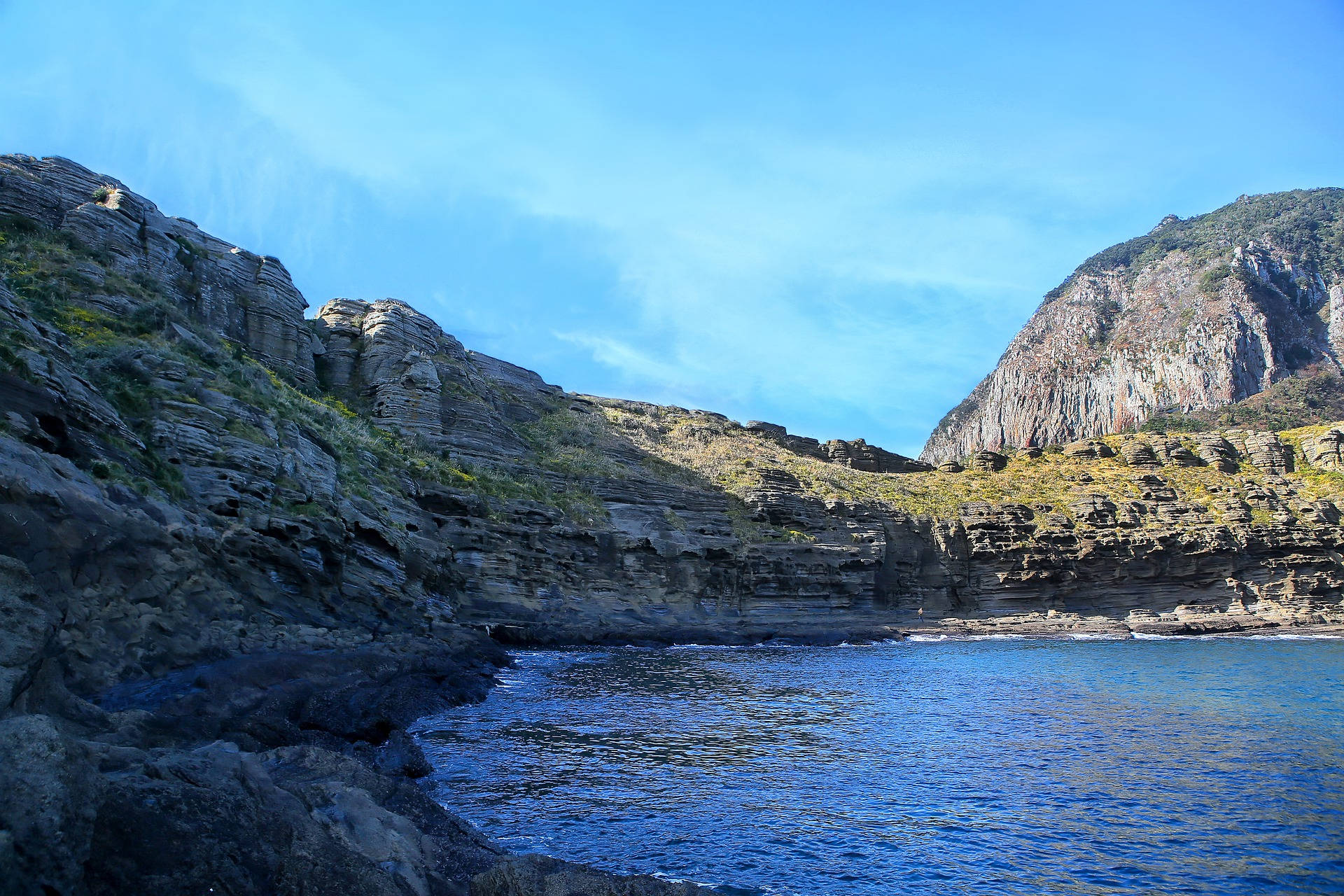 Jeju 桌布 壁紙 — 一個寧靜的海灣，深藍色的海水隱藏在崎嶇的層狀懸崖之間，上方是清澈的藍天。