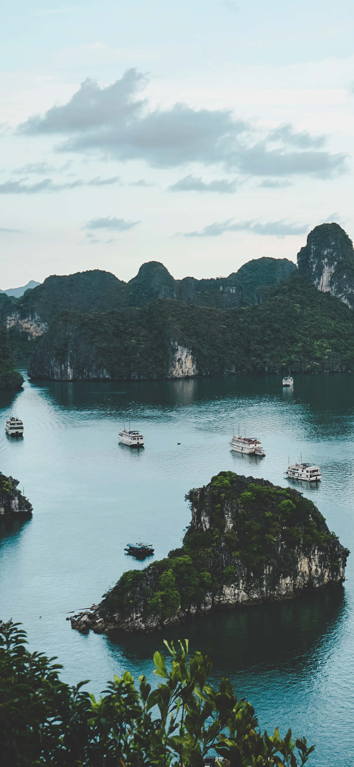 越南 桌布 壁紙 — 一個寧靜的海灣，石灰岩喀斯特地貌從碧綠的海水中升起，海面上點綴著小船，天空多雲。