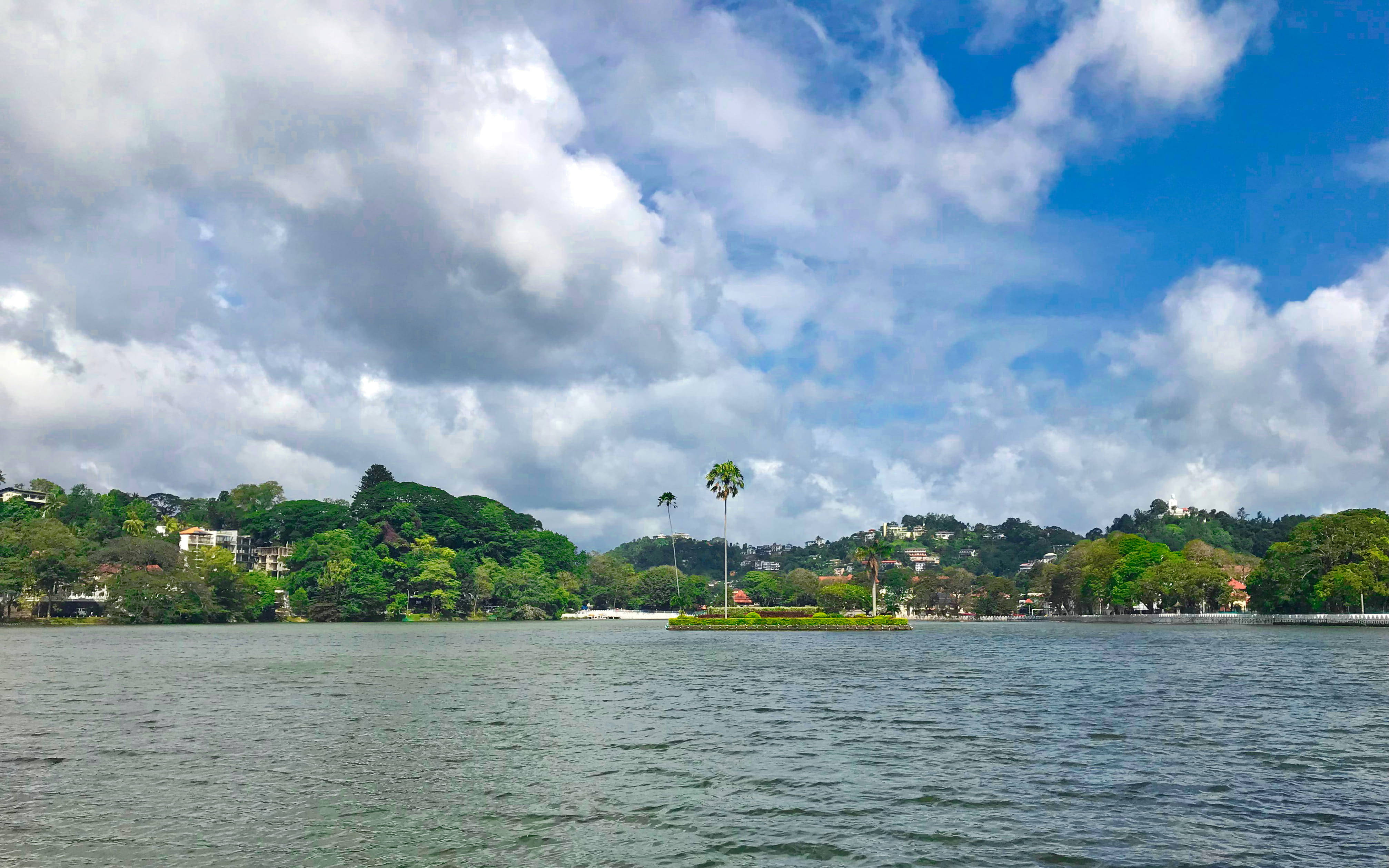 Sri Lanka 桌布 壁紙 — 一個寧靜的湖泊，周圍環繞著茂密的綠色山丘和散落的建築物，天空多雲，藍天白雲。