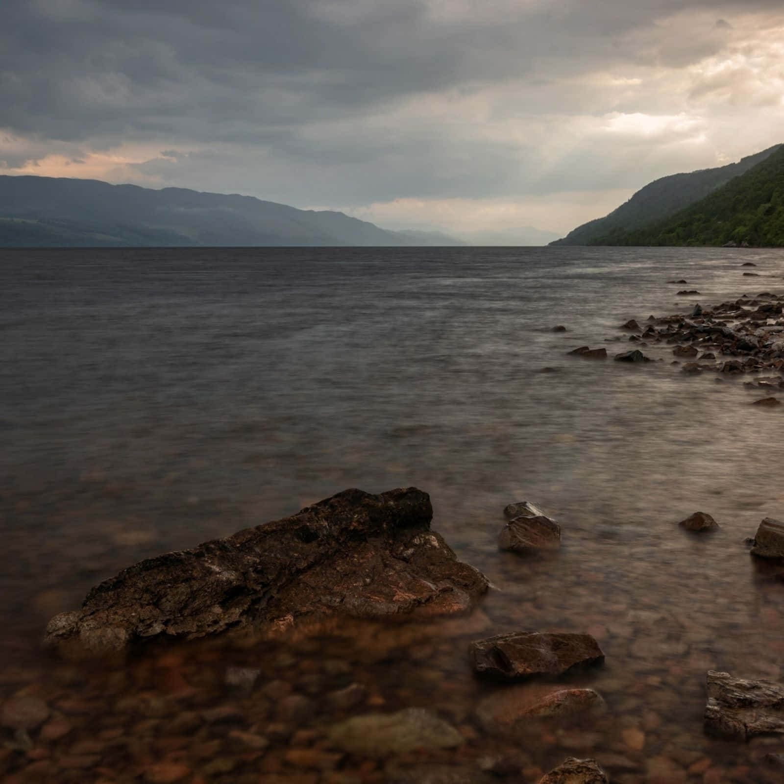 Lake 桌布 壁紙 — 一個寧靜的湖泊，平滑的水流經過岩石海岸，在陰沉多雲的天空下，遠山可見。