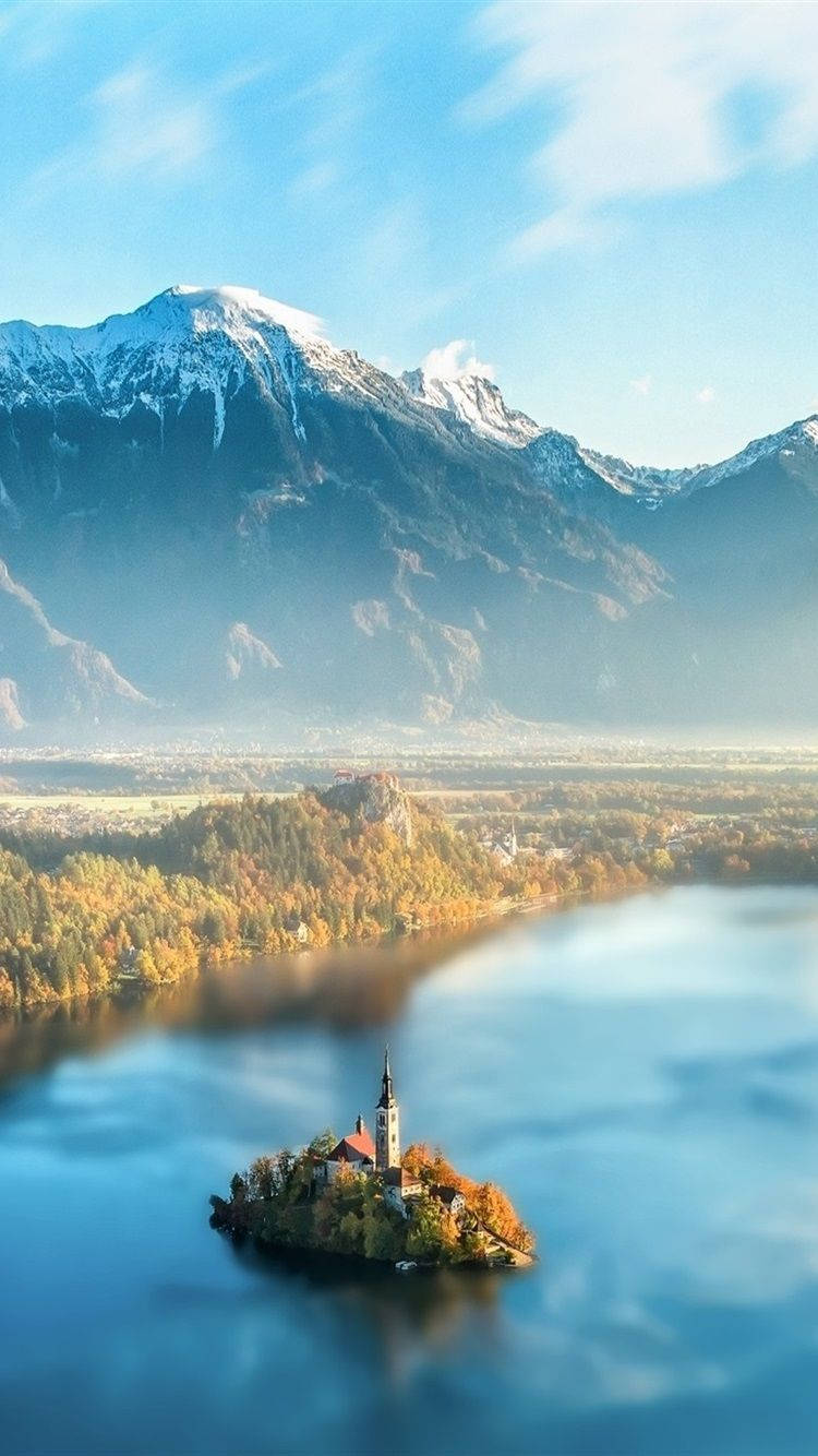 Slovenia 桌布 壁紙 — 一個寧靜的湖泊，湖中小島上有一座教堂，周圍環繞著秋天的樹木和白雪覆蓋的山脈，天空湛藍清澈。