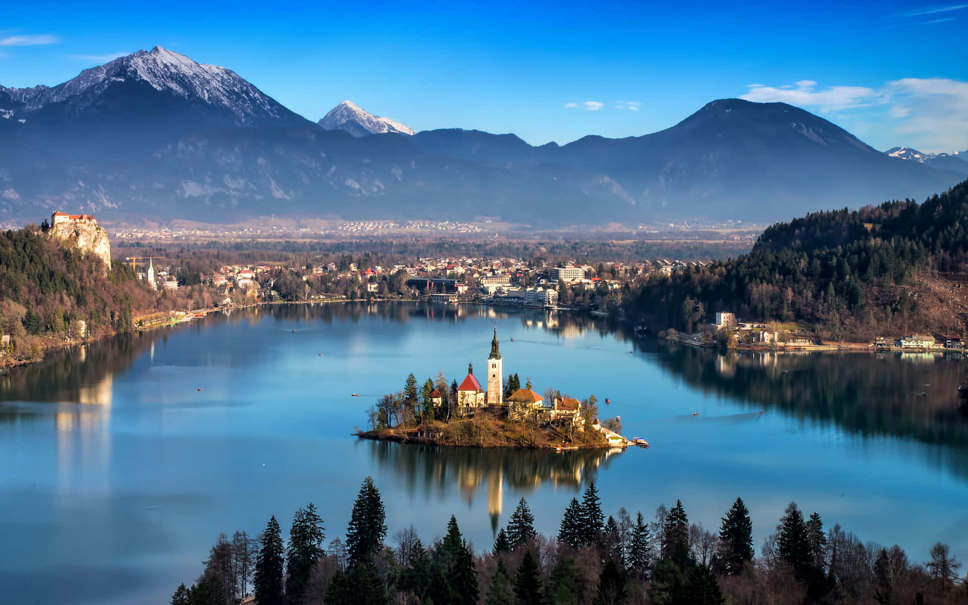 Slovenia 桌布 壁紙 — 一個寧靜的湖泊，湖中有一個小島教堂，周圍是白雪覆蓋的山脈和樹木繁茂的海岸線，在清澈的藍天下。