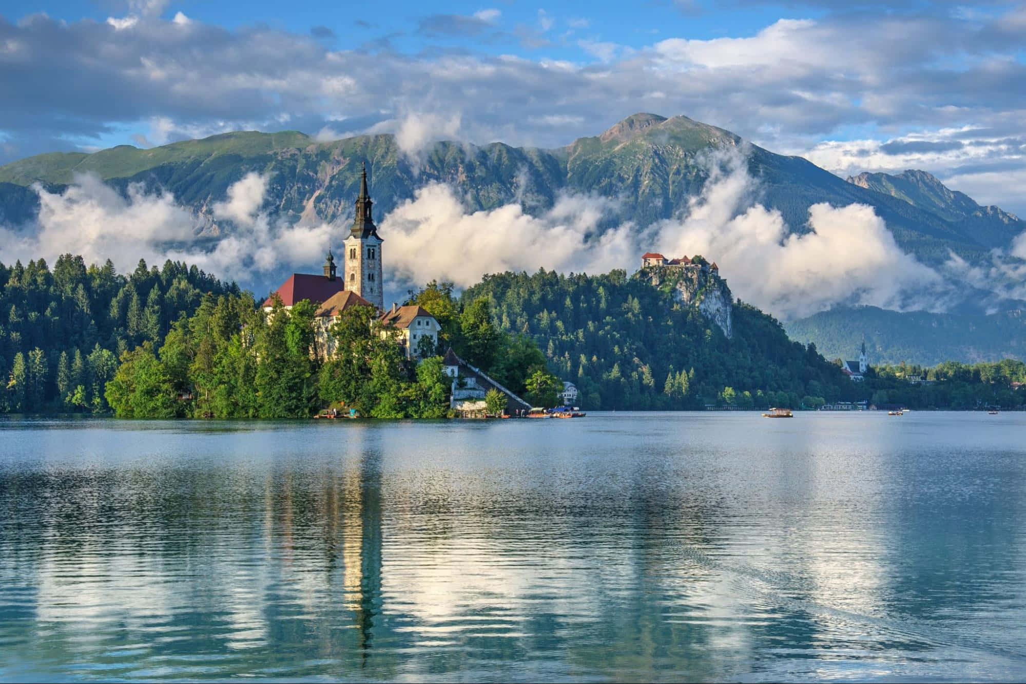 Alps 桌布 壁紙 — 一個寧靜的湖泊倒映著一座歷史悠久的教堂，教堂坐落於一個鬱鬱蔥蔥的島嶼上，周圍環繞著煙霧繚繞的山脈，天空部分多雲。