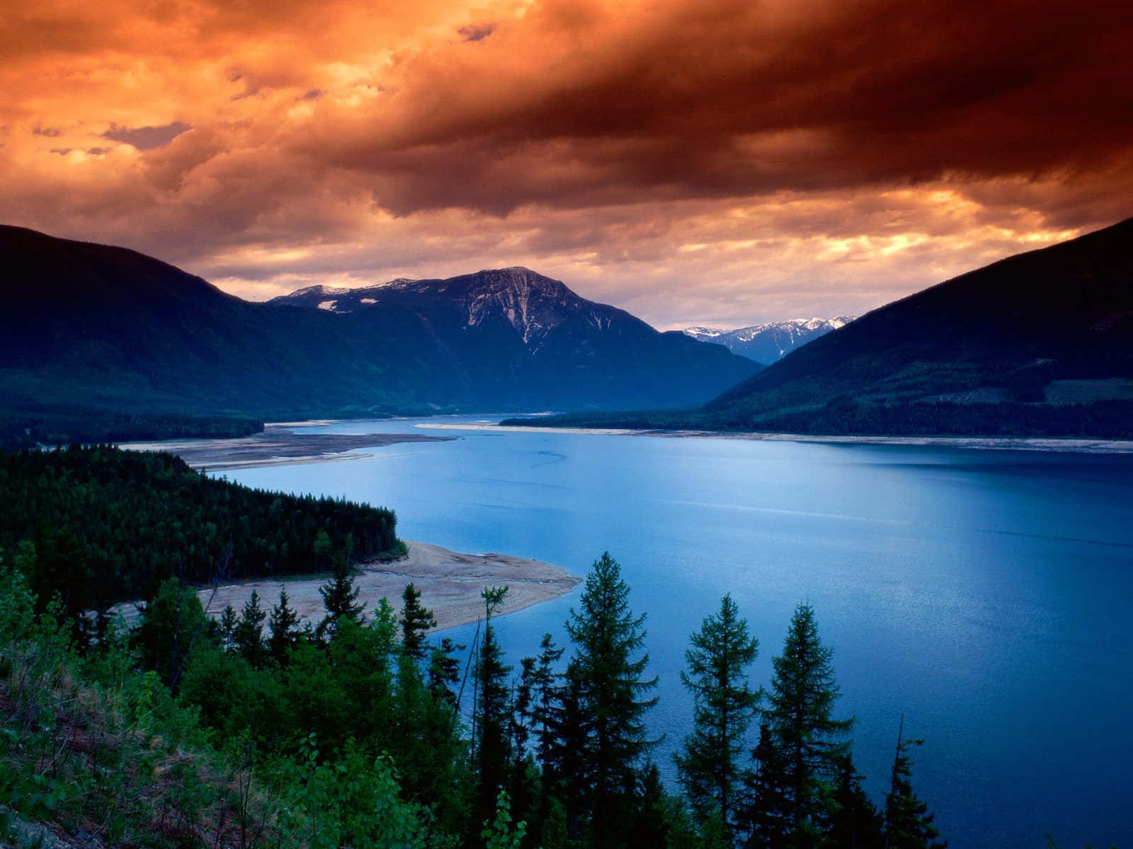 Beautiful Lake 桌布 壁紙 — 一個寧靜的湖泊坐落在森林覆蓋的山脈之間，在火紅的橙色日落天空下。