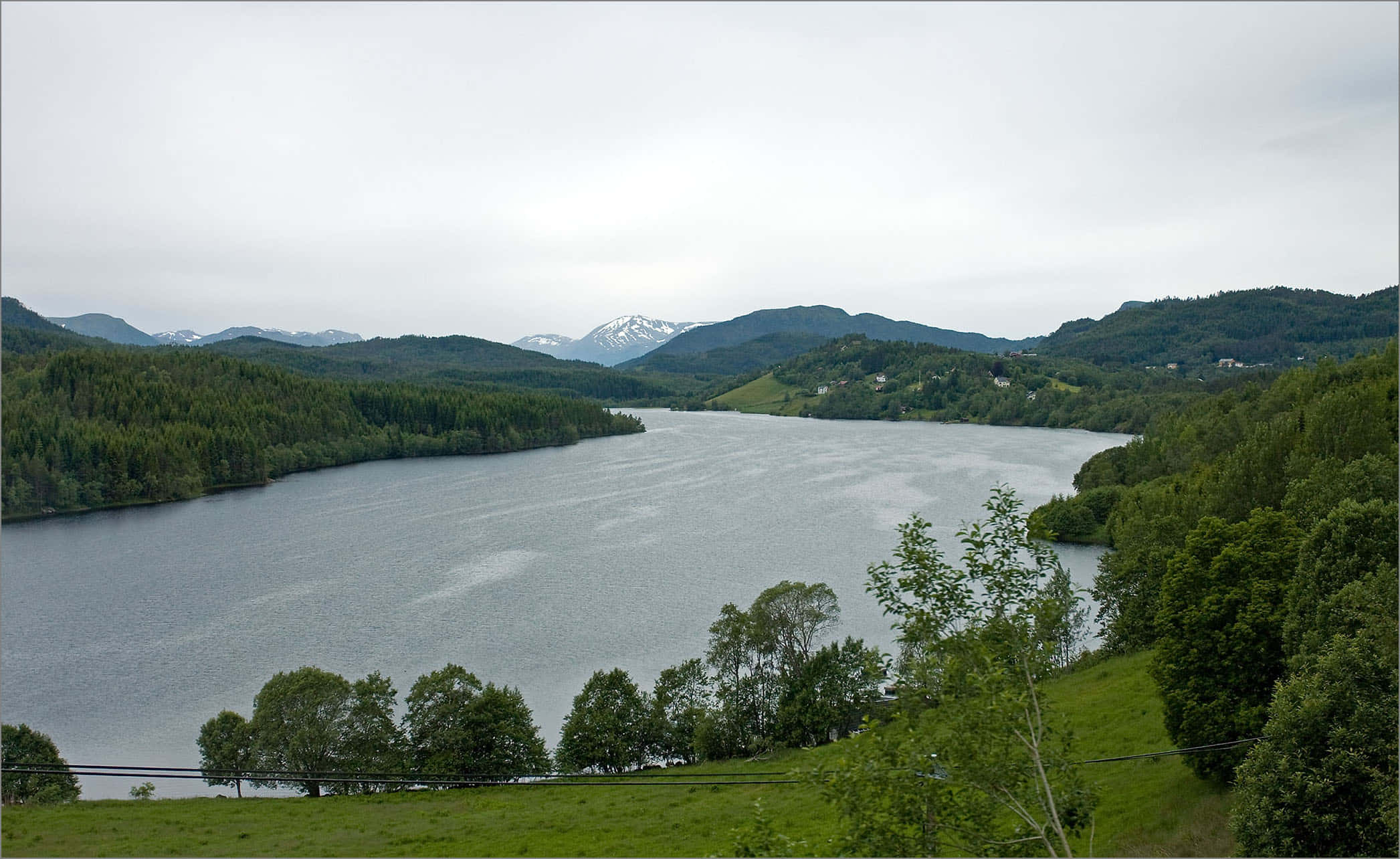 Lake 桌布 壁紙 — 一個寧靜的湖泊坐落在鬱鬱蔥蔥的青山之間，遠處可見白雪覆蓋的山峰，天空多雲。