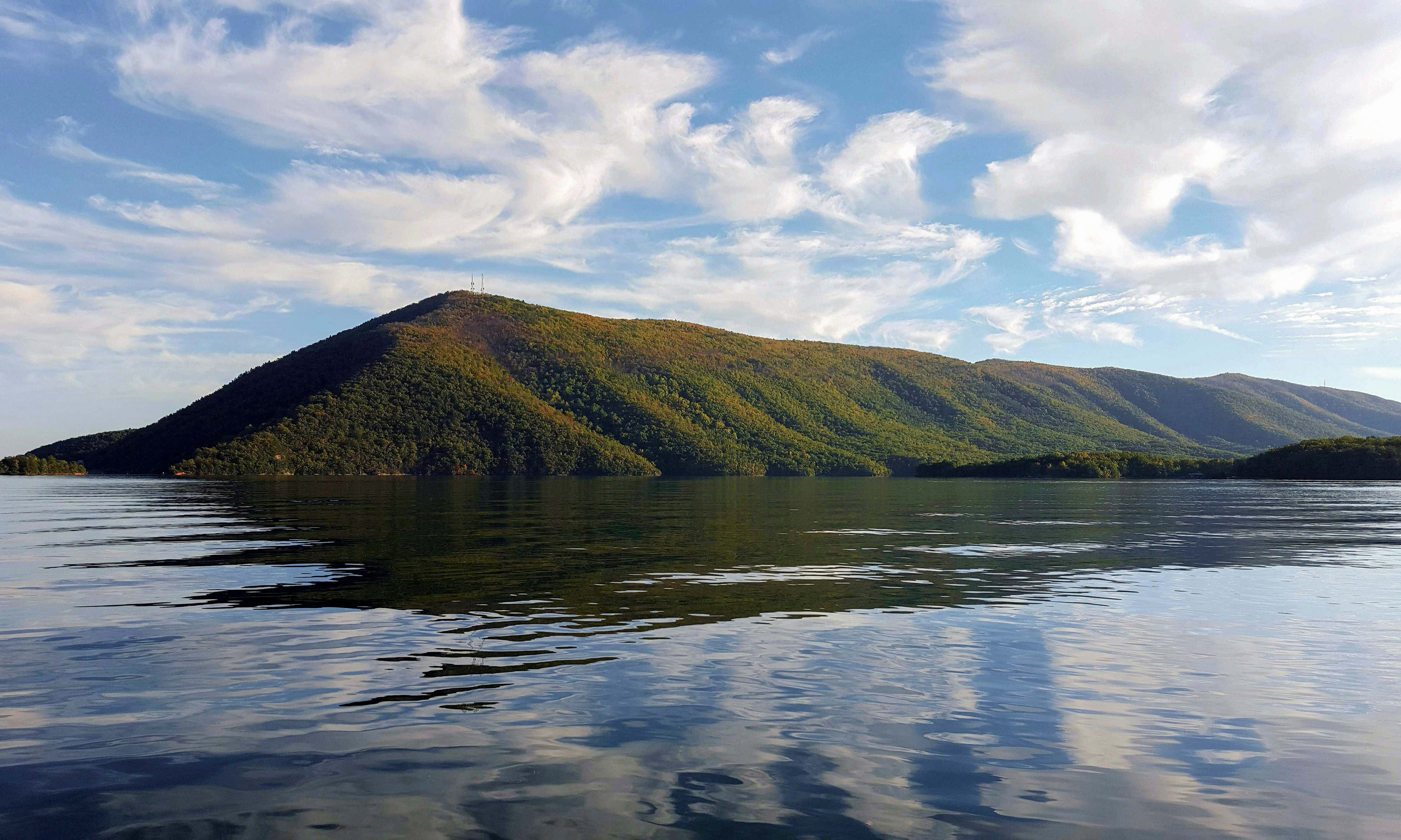 Lake 桌布 壁紙 — 一個寧靜的湖泊映照著一片樹林覆蓋的山丘，天空湛藍，飄著幾縷輕雲。