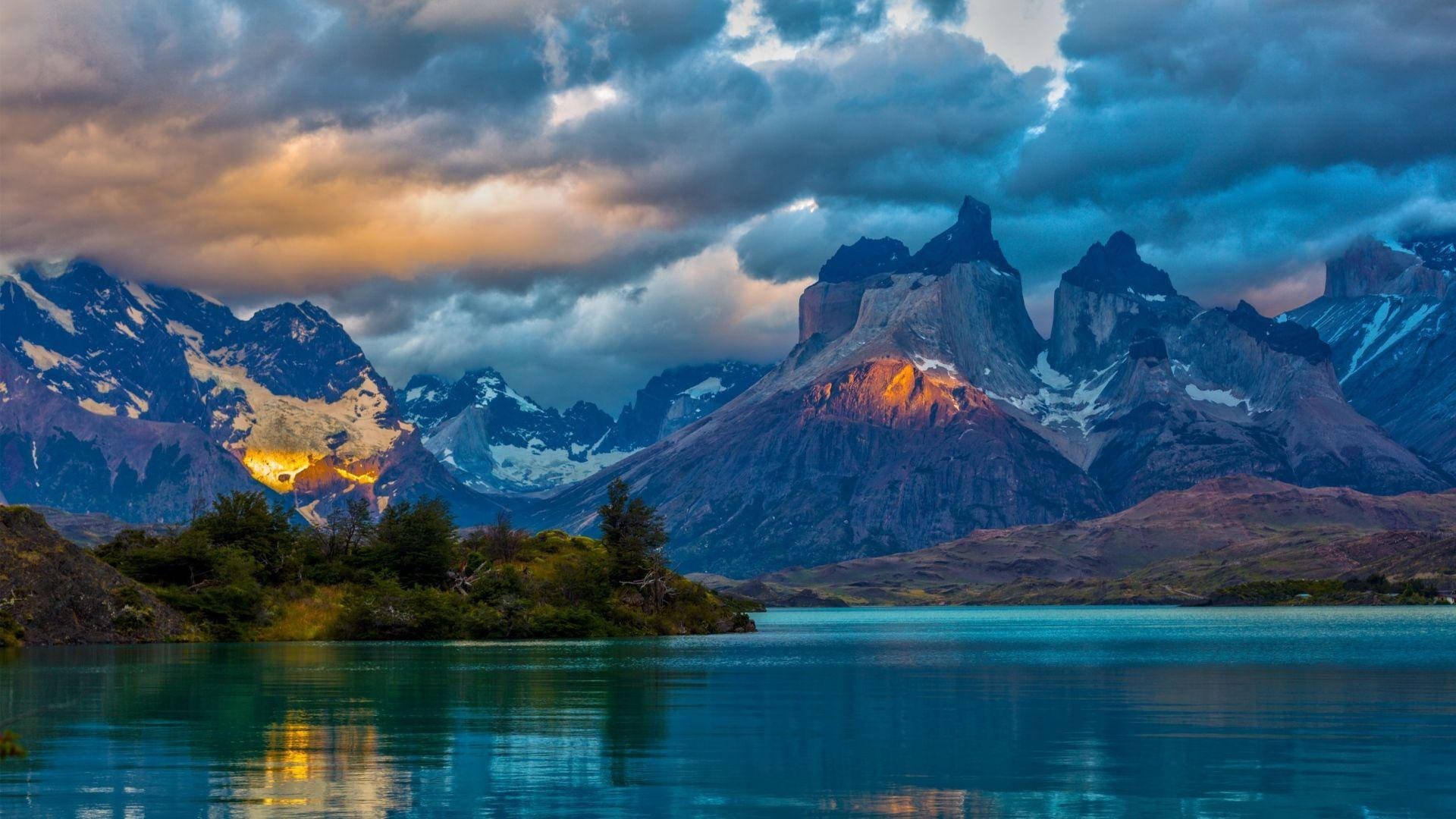 Patagonia 桌布 壁紙 — 一個寧靜的湖泊映照著在戲劇性的雲層下，被金色夕陽照亮、積雪覆蓋的山峰。