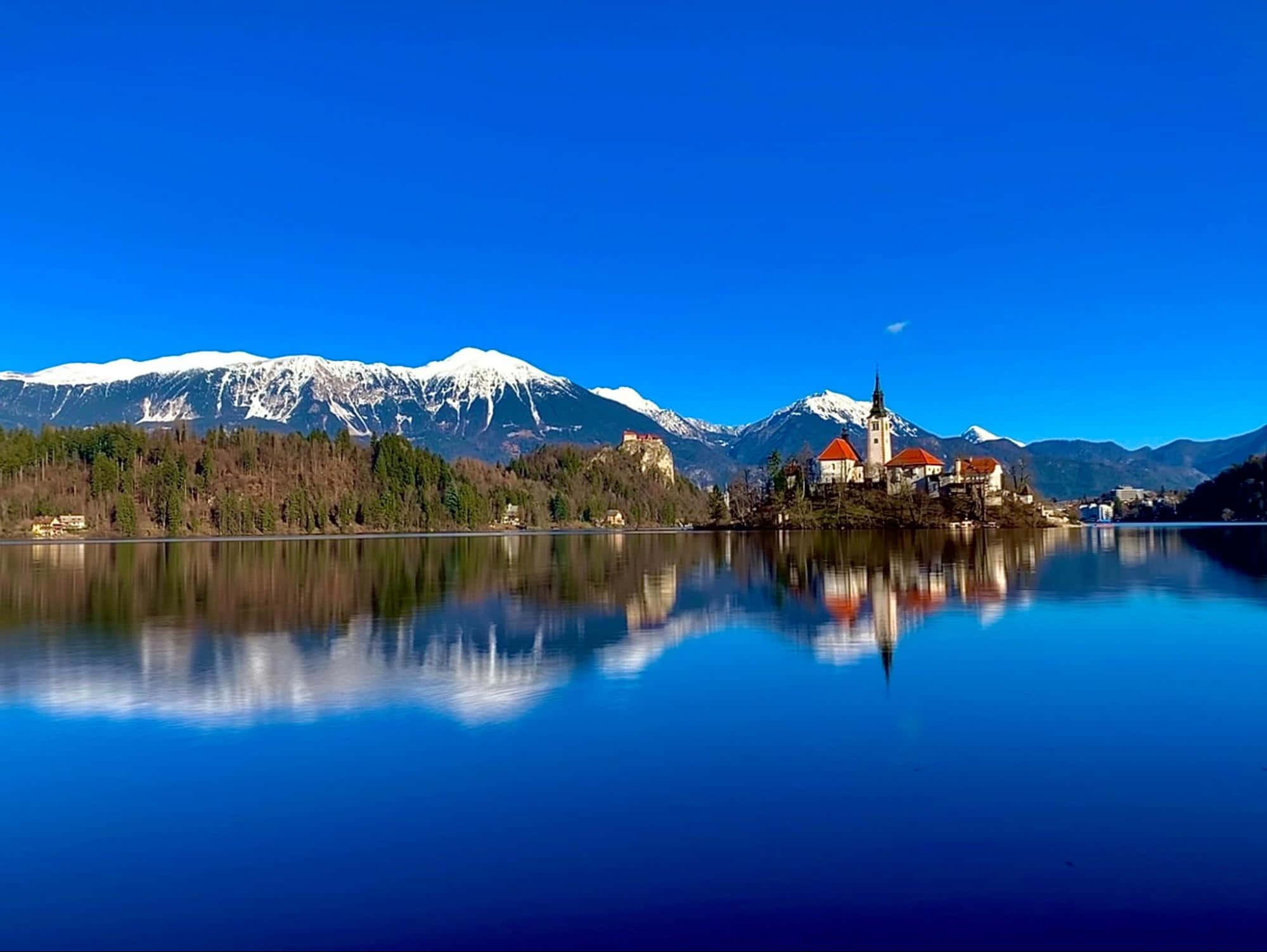 Alps 桌布 壁紙 — 一個寧靜的湖泊映照著白雪覆蓋的山脈和一個小島上紅頂教堂，在清澈的藍天下。