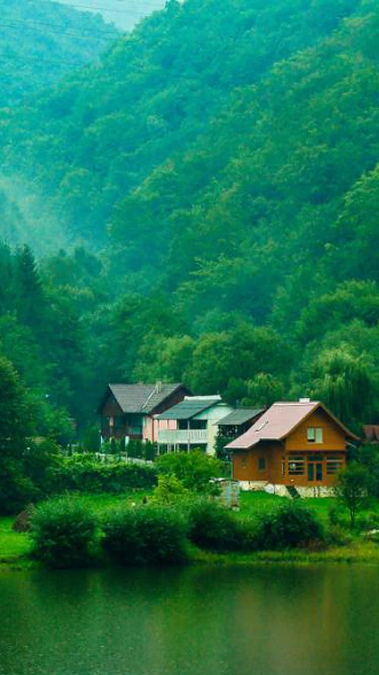 Green Nature iPhone 桌布 壁紙 — 一個寧靜的湖邊村莊，坐落在鬱鬱蔥蔥的綠色山脈中，背景是霧氣瀰漫的森林山丘。