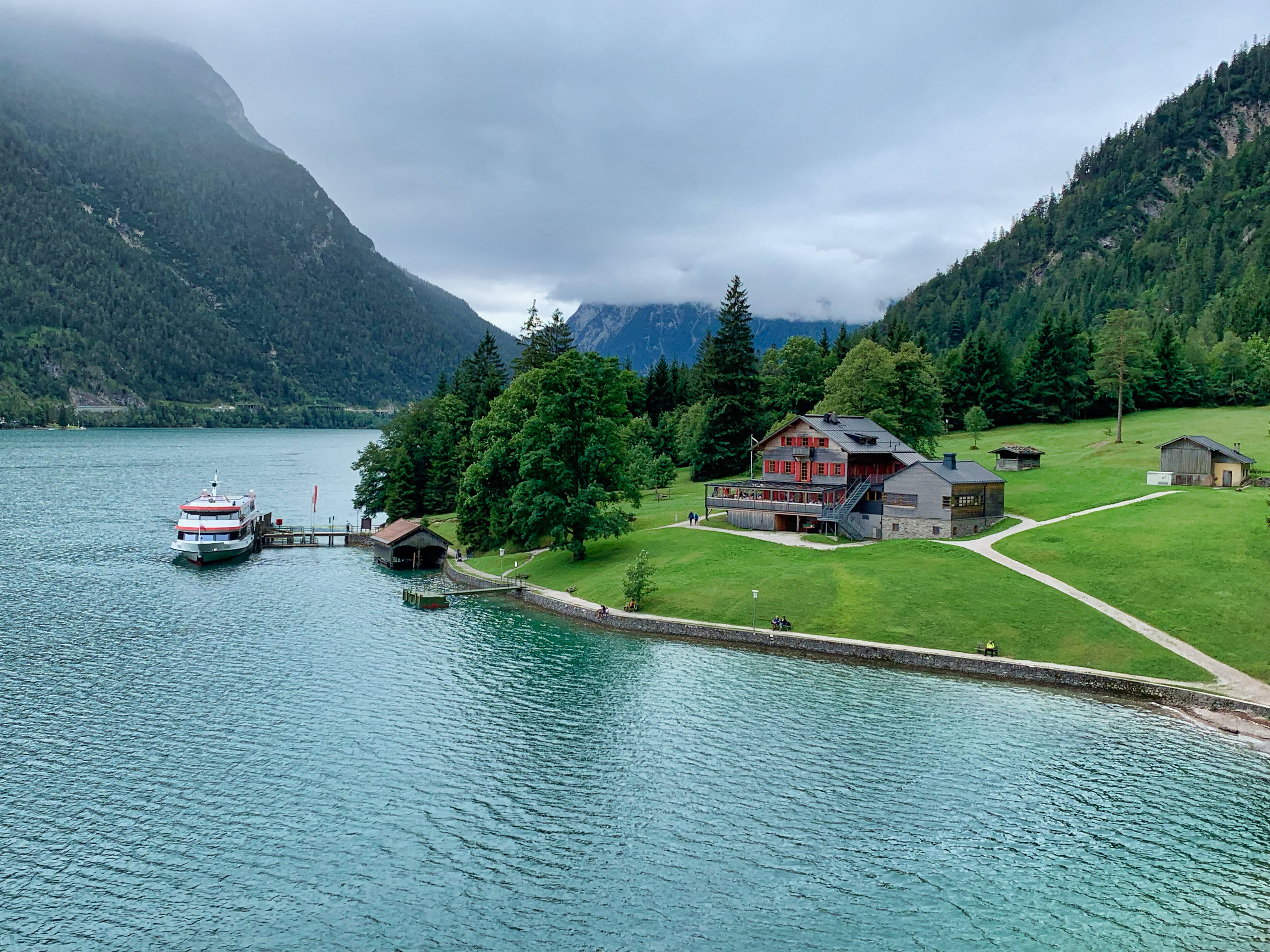 Austria 桌布 壁紙 — 一個寧靜的湖邊村莊，紅頂小木屋坐落在綠色的山丘和高聳的森林覆蓋的山脈之間，天空多雲。