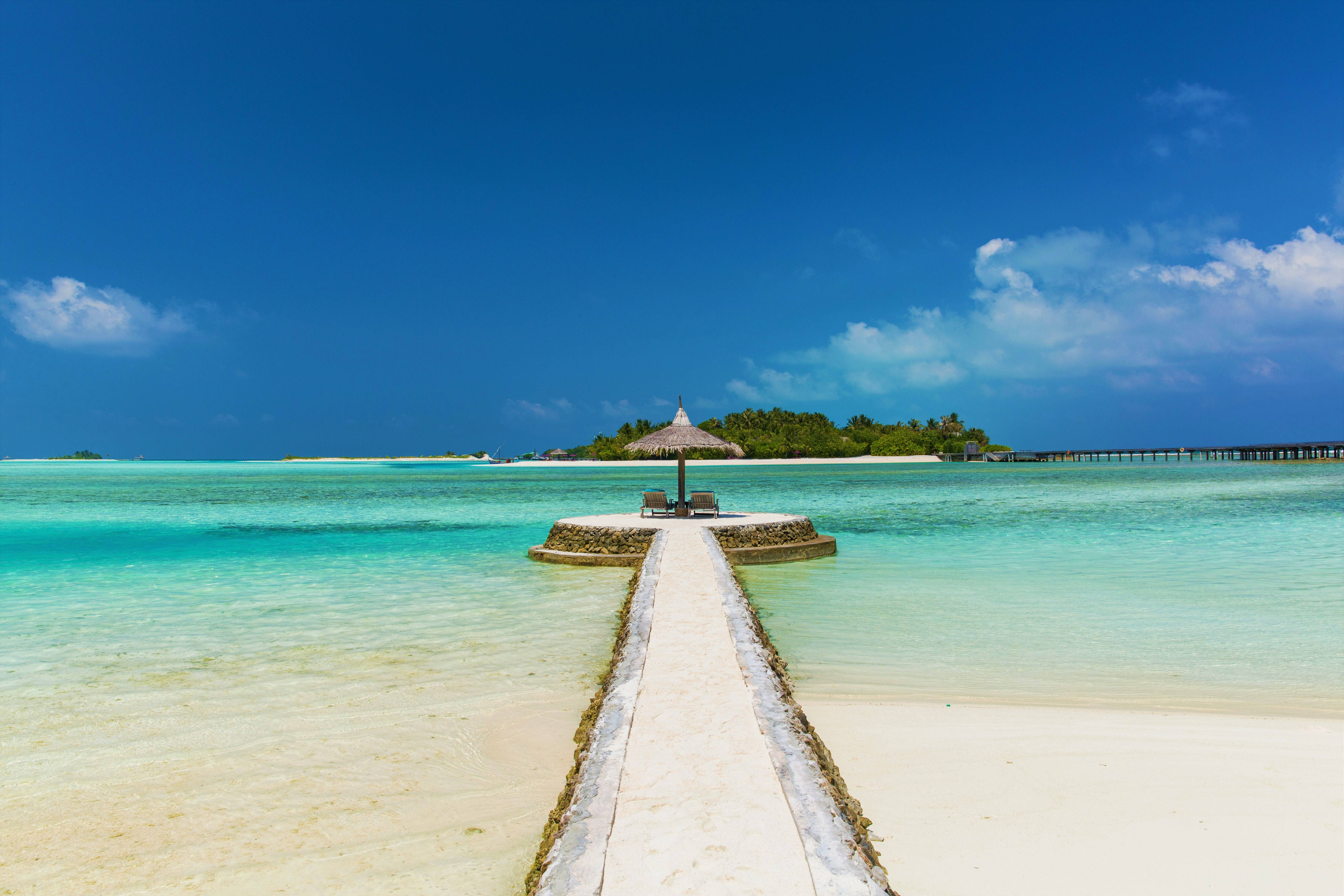 Maldives 桌布 壁紙 — 一個寧靜的熱帶島嶼，擁有碧綠的海水，以及從白色沙灘延伸出的石碼頭上的茅草屋頂涼亭。