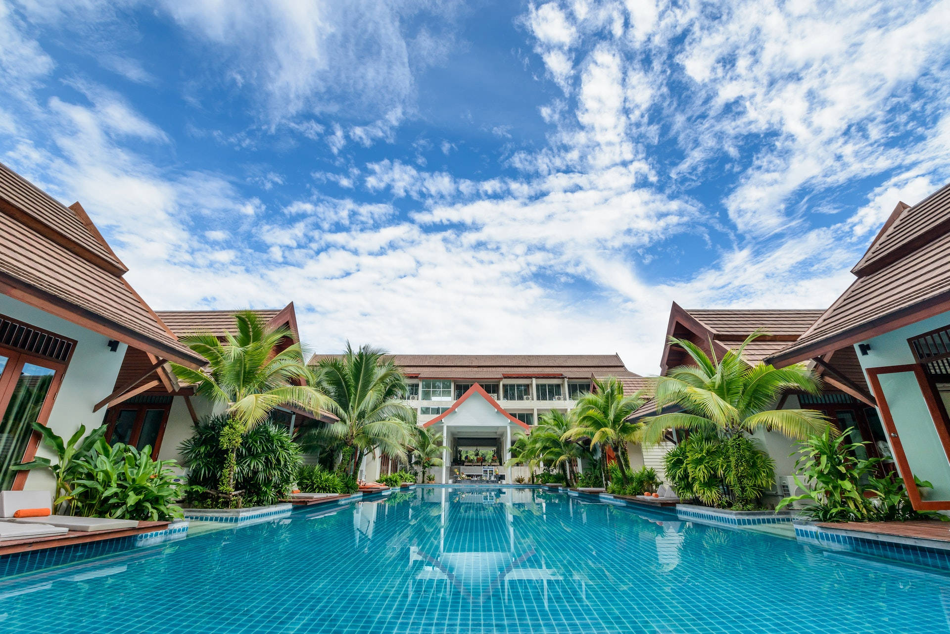Hotel 桌布 壁紙 — 一個寧靜的熱帶度假村泳池，兩旁種滿棕櫚樹和傳統屋頂別墅，在明亮的藍天下有縷縷白雲。