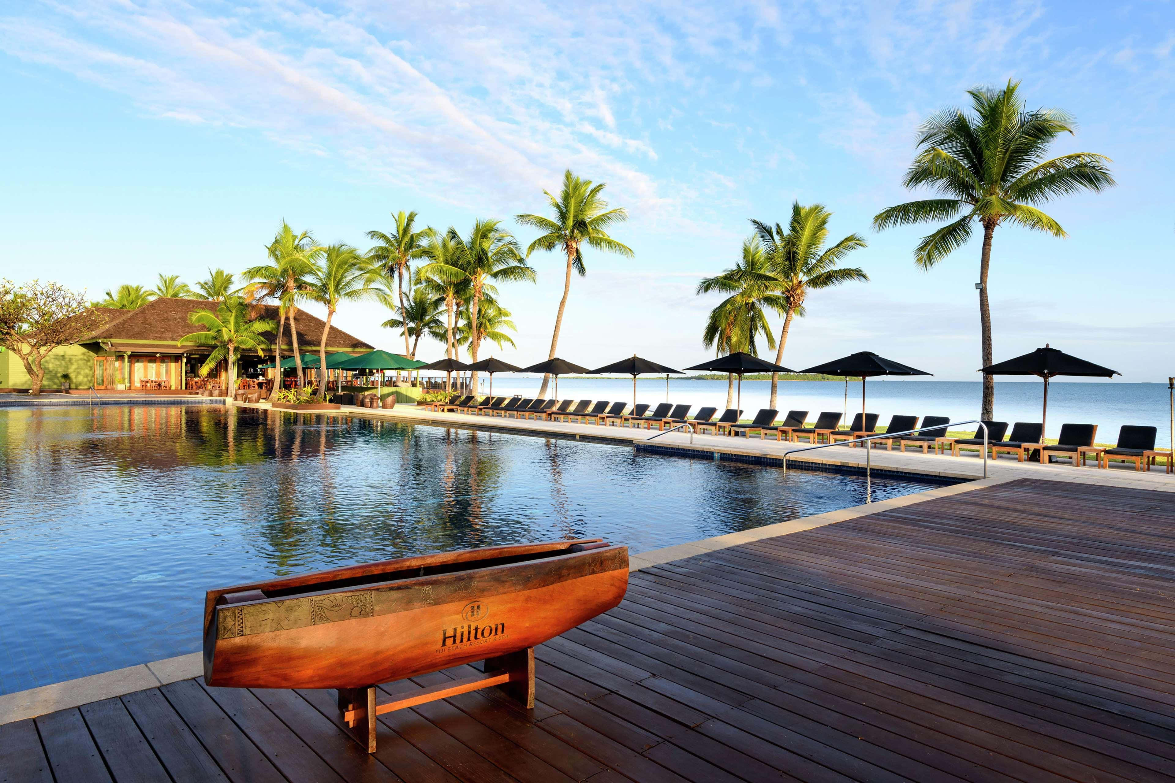 Fiji Beach 桌布 壁紙 — 一個寧靜的熱帶度假村泳池，棕櫚樹和躺椅，在清澈的藍天下俯瞰海洋。