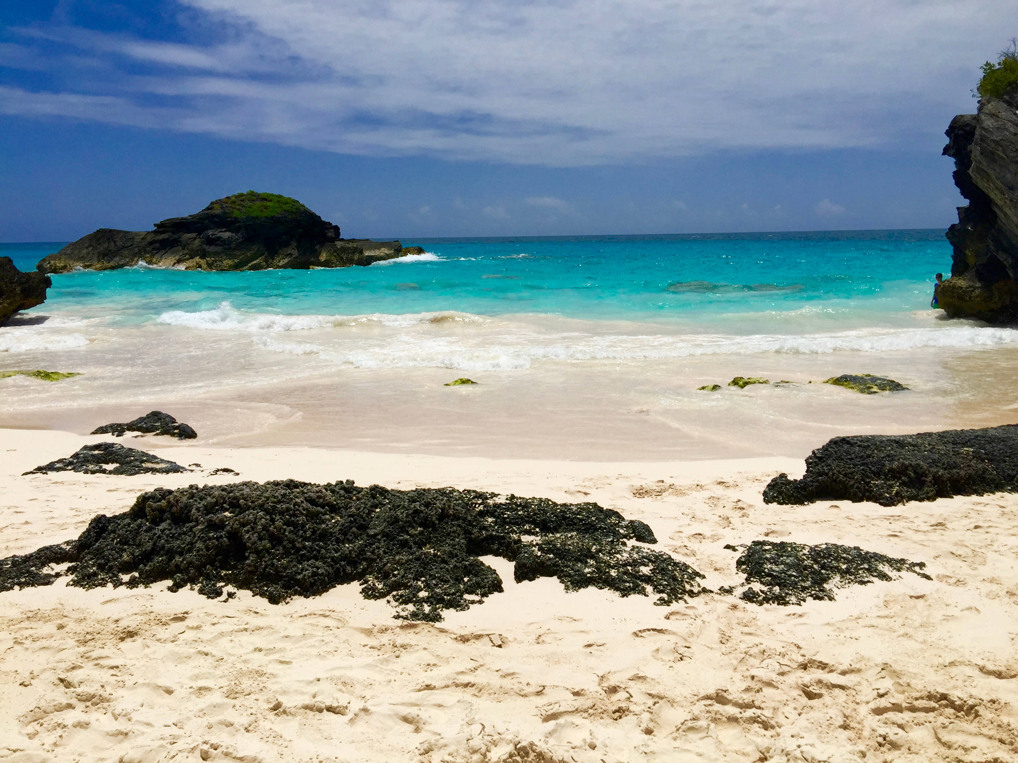 Bermuda 桌布 壁紙 — 一個寧靜的熱帶海灘，碧綠的海水拍打著白色沙灘，周圍是深色的火山岩和茂密的綠色懸崖，天空部分多雲。