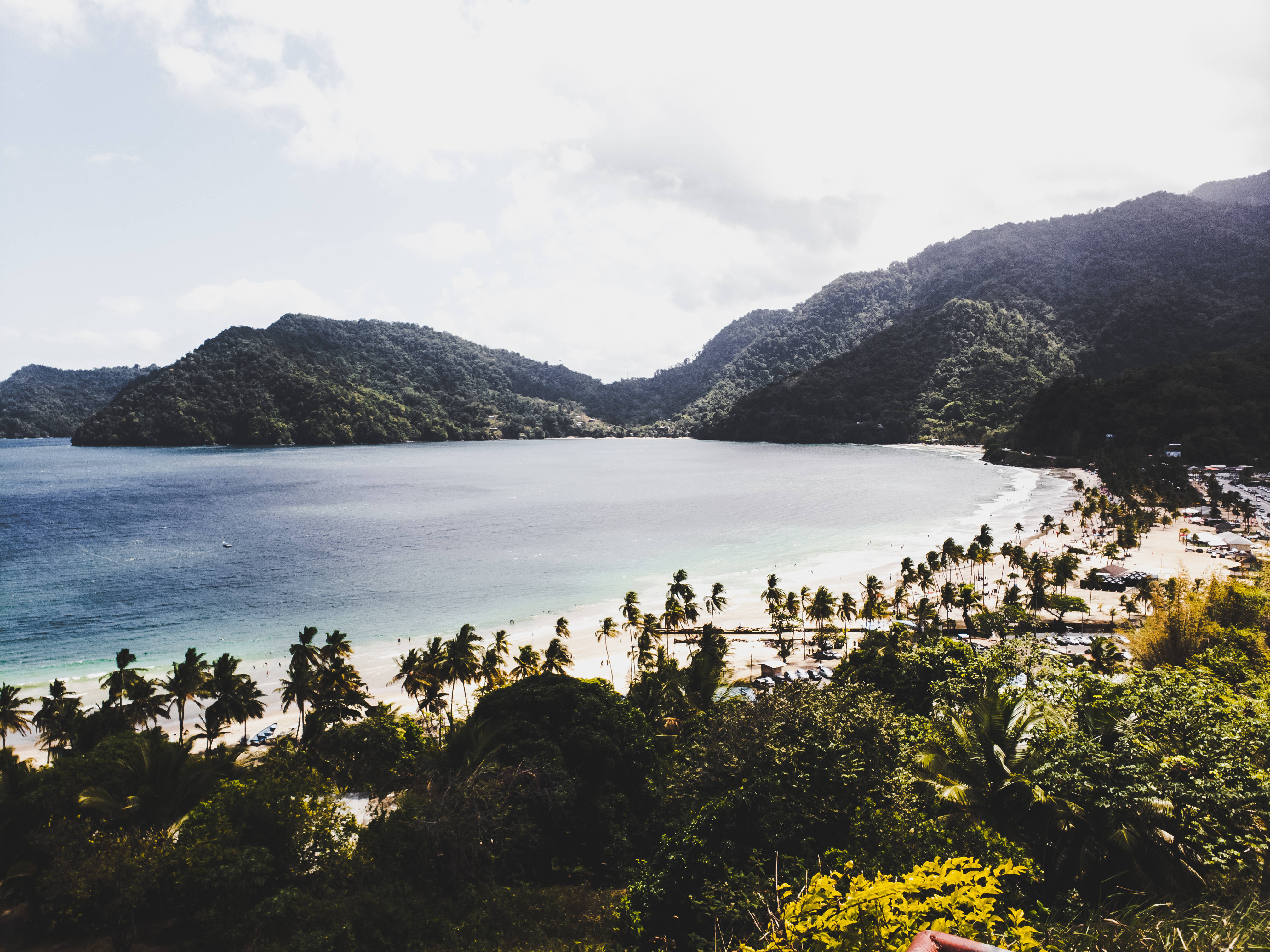 Trinidad 桌布 壁紙 — 一個寧靜的熱帶海灣，被茂密的綠色山脈環繞，沙灘上排列著棕櫚樹，天空明亮。